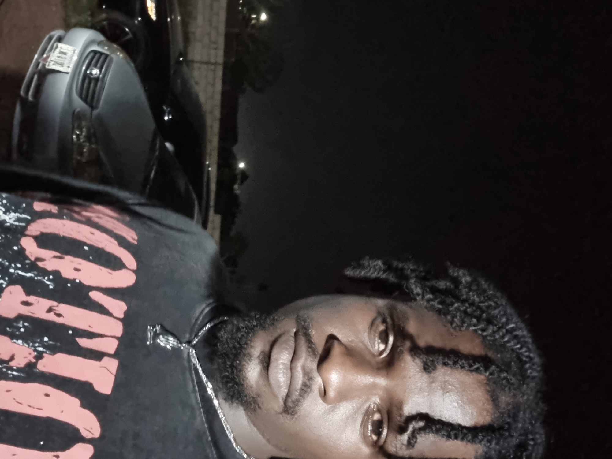 A man with dark skin, short, twisted black hair, and a goatee stands outdoors at night. He wears a black t-shirt with a light-colored graphic design and a silver chain around his neck. Behind him, a silver car is partially visible, and further back, there's a brick wall and what appears to be street lighting. The location is Bar Jirgi Summa, Nigeria.