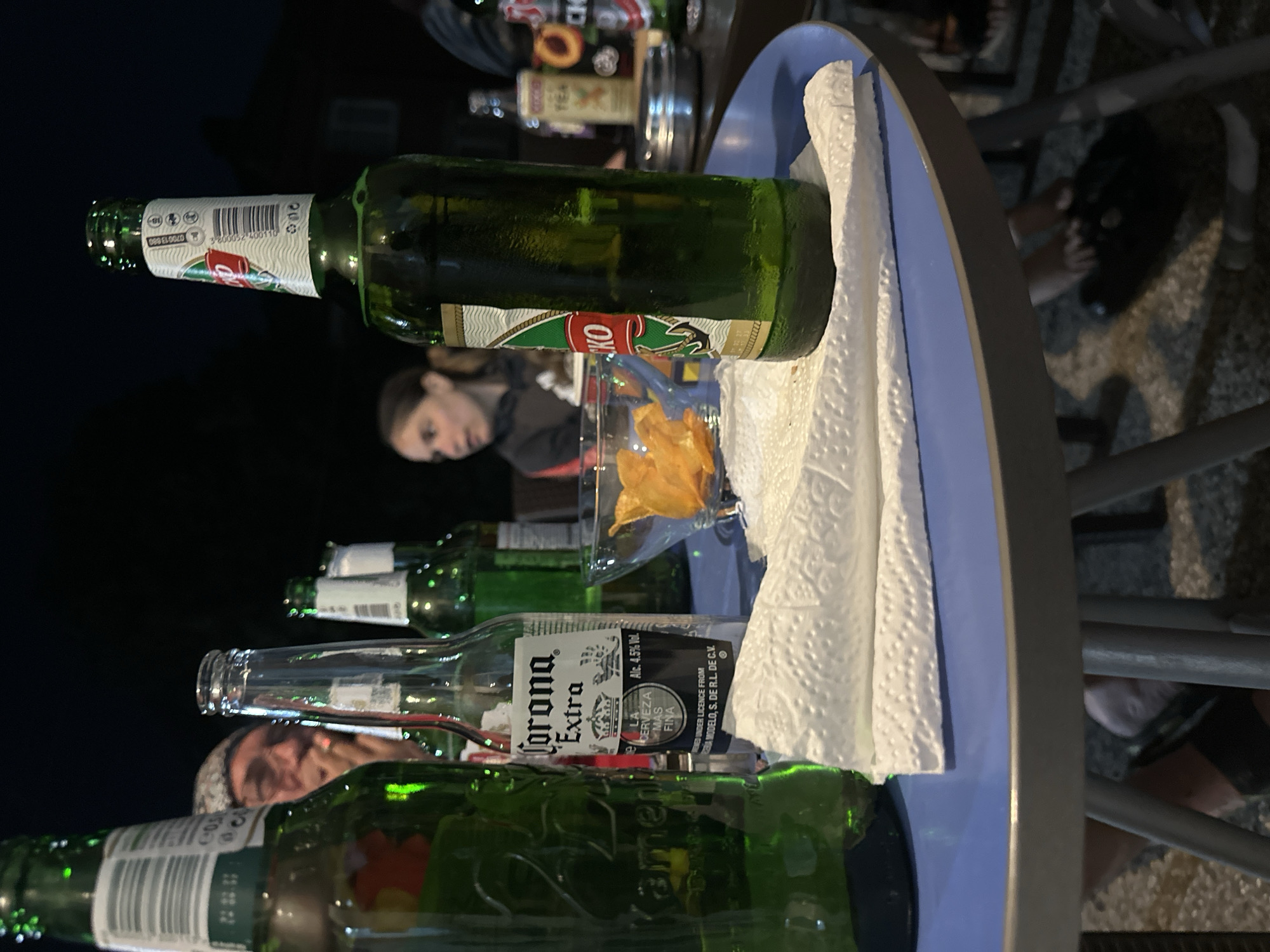 This is a low-angle, close-up, outdoor nighttime photograph of a table set with several beer bottles and a bowl of chips. In the foreground, to the left, is a large green beer bottle, partially obscuring the view. To its right, a clear glass bottle with a "Corona Extra" label is visible. Further to the right, a green bottle with a red and white label is prominently featured in the center. A glass bowl filled with orange potato chips sits behind the Corona bottle. In the background, a man is partially visible, smoking a cigarette and looking towards the left. A young woman is also visible in the background, facing towards the left, wearing a dark hooded sweatshirt. The table surface is blue. The scene appears to be an informal gathering. The location is Ekzarh Antimovo, Bulgaria.