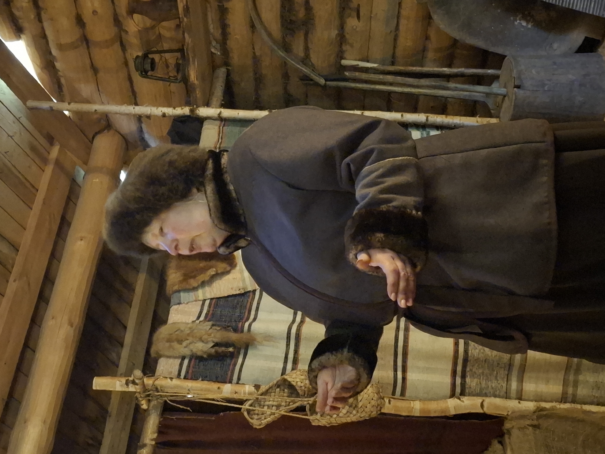 The image shows a woman wearing a dark, fur-trimmed coat and hat. She is standing in a room with a wooden beam ceiling. There is a woven rug and a pair of woven shoes on a shelf in front of her. A lantern hangs from the ceiling. The location is Unknown, Unknown.