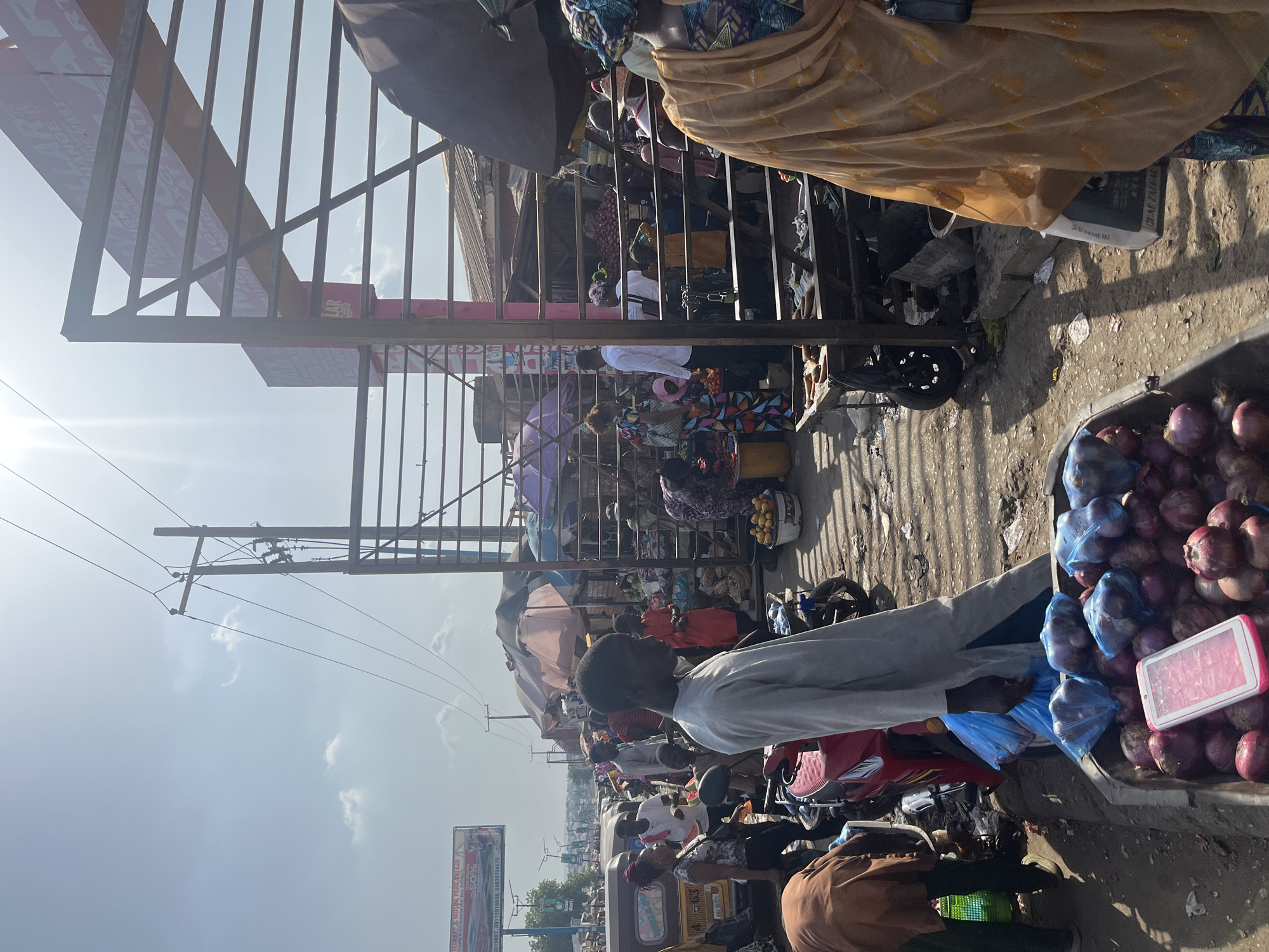 The image depicts a bustling street scene in Minna, Nigeria. People are crowded together amidst vendors, vehicles (including motorcycles and tuk-tuks), and market stalls. A man in a light-colored shirt is positioned in the foreground next to a vendor selling onions in blue plastic bags. Overhead, a metal structure provides some shade, and power lines stretch across the sky. In the background, a billboard is visible. The scene is chaotic and lively, characteristic of a busy market environment.