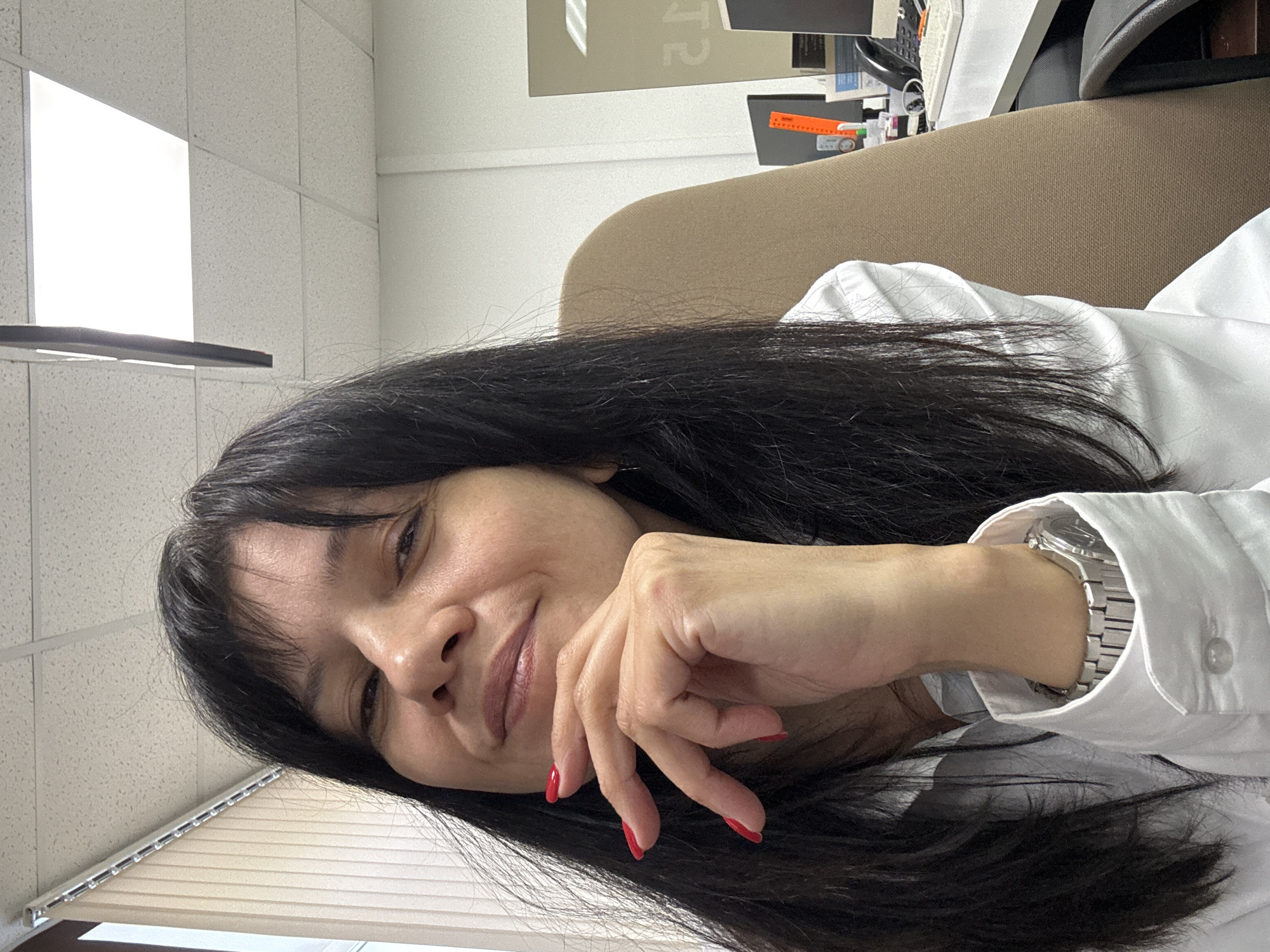 A woman with long, dark hair, red nail polish, and a silver wristwatch is seen in a selfie-style image. She is wearing a white collared shirt. Her face is slightly turned and tilted. Behind her is what appears to be an office setting, including a window with blinds, a ceiling with tile-like squares, a beige office chair, and desk items. The location is Chaikovsky, Russia.