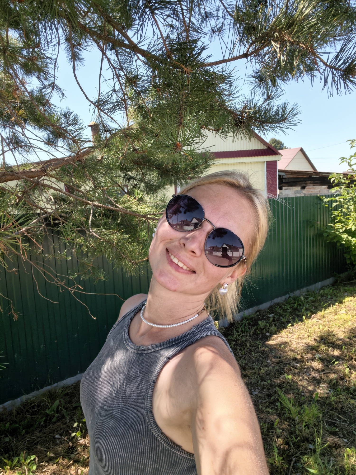 A blonde woman with fair skin, wearing round sunglasses, a grey tank top, and a pearl necklace, smiles and poses for a selfie outdoors. She has a pearl earring in her left ear. The background features a green corrugated metal fence, a pine tree branch in the foreground, and houses with red and yellow roofs. The scene appears to be a residential area in Sterlitamak, Russia, under a clear blue sky. Sunlight casts shadows on the ground and on the woman's clothing.