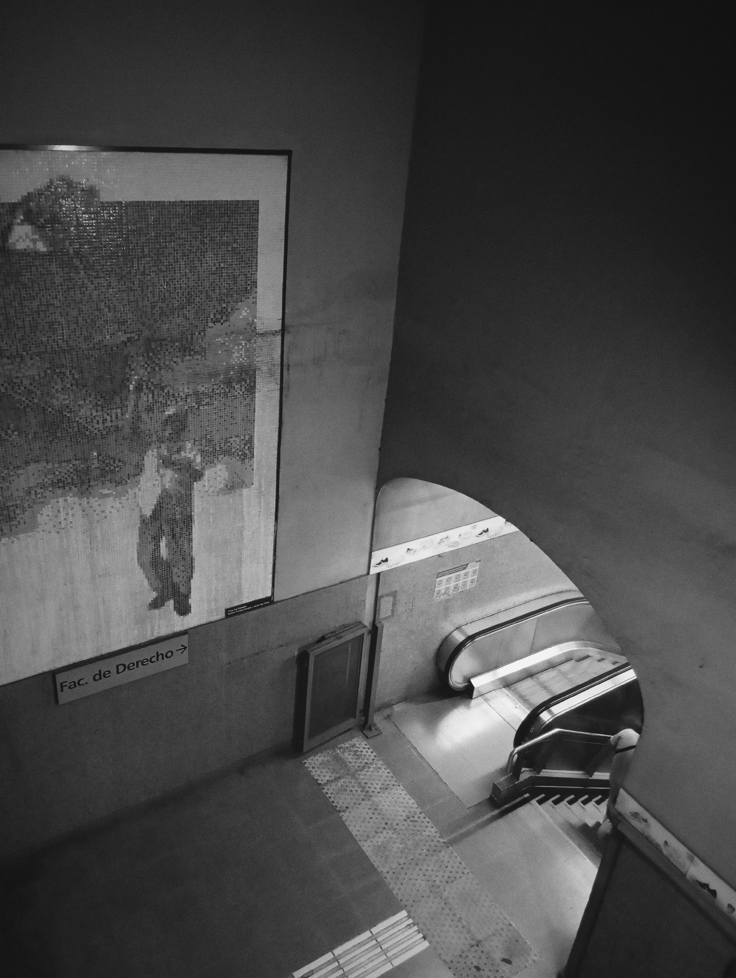 This black and white image captures an interior section of the Facultad de Derecho - Julieta Lanteri subway station on the H line in Buenos Aires, Argentina, viewed from an elevated perspective. A large, framed mosaic artwork is mounted on the upper left wall, depicting a stylized human figure, possibly male, holding an object. Below the mosaic, a white rectangular sign with a right-pointing arrow reads "Fac. de Derecho". The floor in the foreground consists of plain grey paving transitioning into a section of tactile textured tiles. To the right, an arched passageway opens to reveal a descending escalator with a metal handrail. A person's head and upper back, facing away from the camera, are partially visible at the bottom of the escalator, indicating their presence on the lower platform.