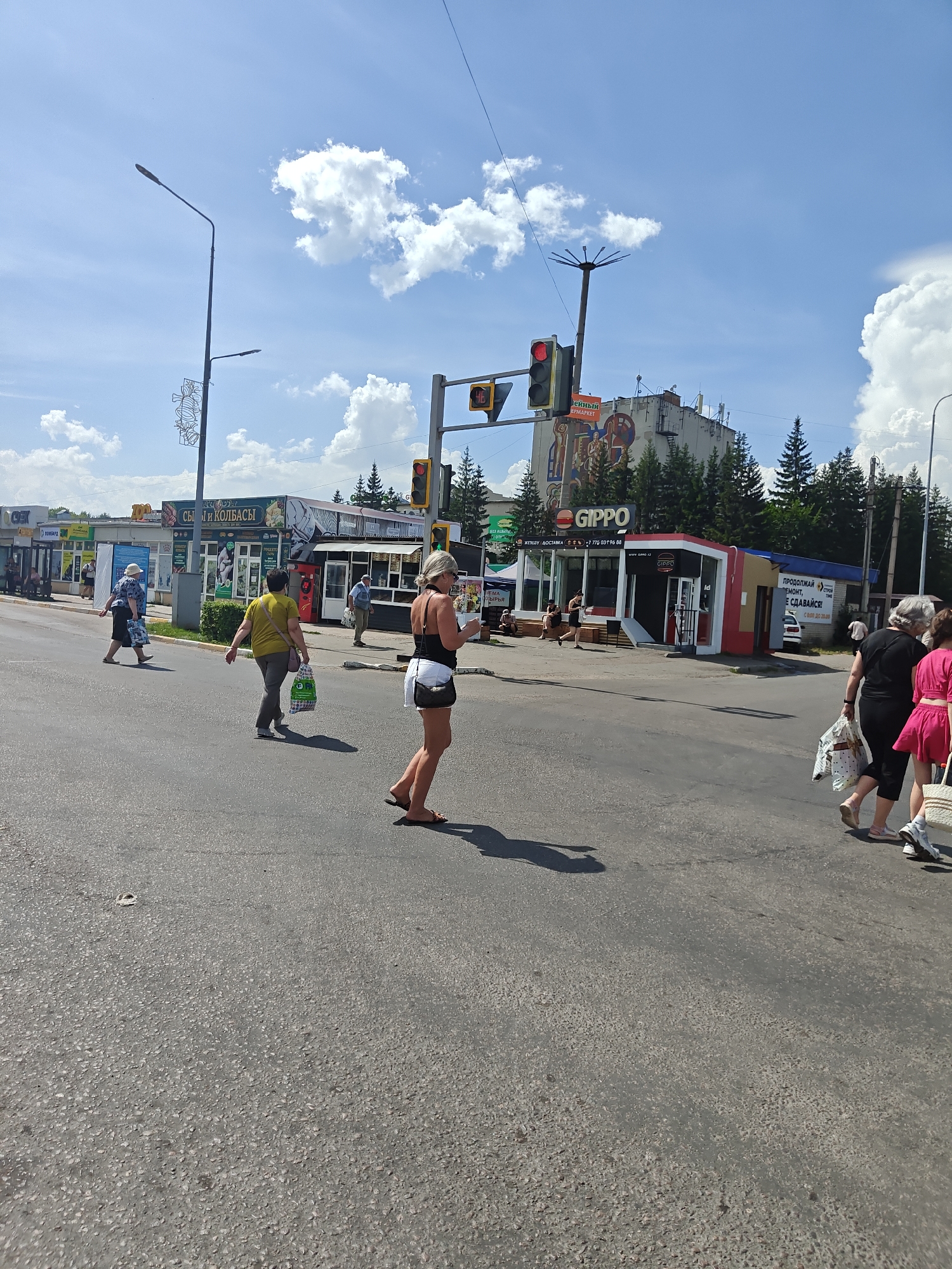 In Petropavl, Kazakhstan, on a sunny day, several people are crossing a road at a pedestrian crossing. A woman in the foreground is walking from right to left, wearing a white skirt and a black tank top, and carrying a handbag. To her left, a woman in a yellow shirt and dark pants is also crossing, holding a shopping bag. Further left, another woman in a blue shirt and dark skirt is walking in the same direction, carrying a shopping bag. In the background, there are several shops and buildings, including a "GIPPO" supermarket and a shop labeled "СЫРЫ И КОЛБАСЫ" (Cheeses and Sausages). Traffic lights are visible at the intersection, and a street lamp stands to the left. The environment appears to be a commercial area within the city.