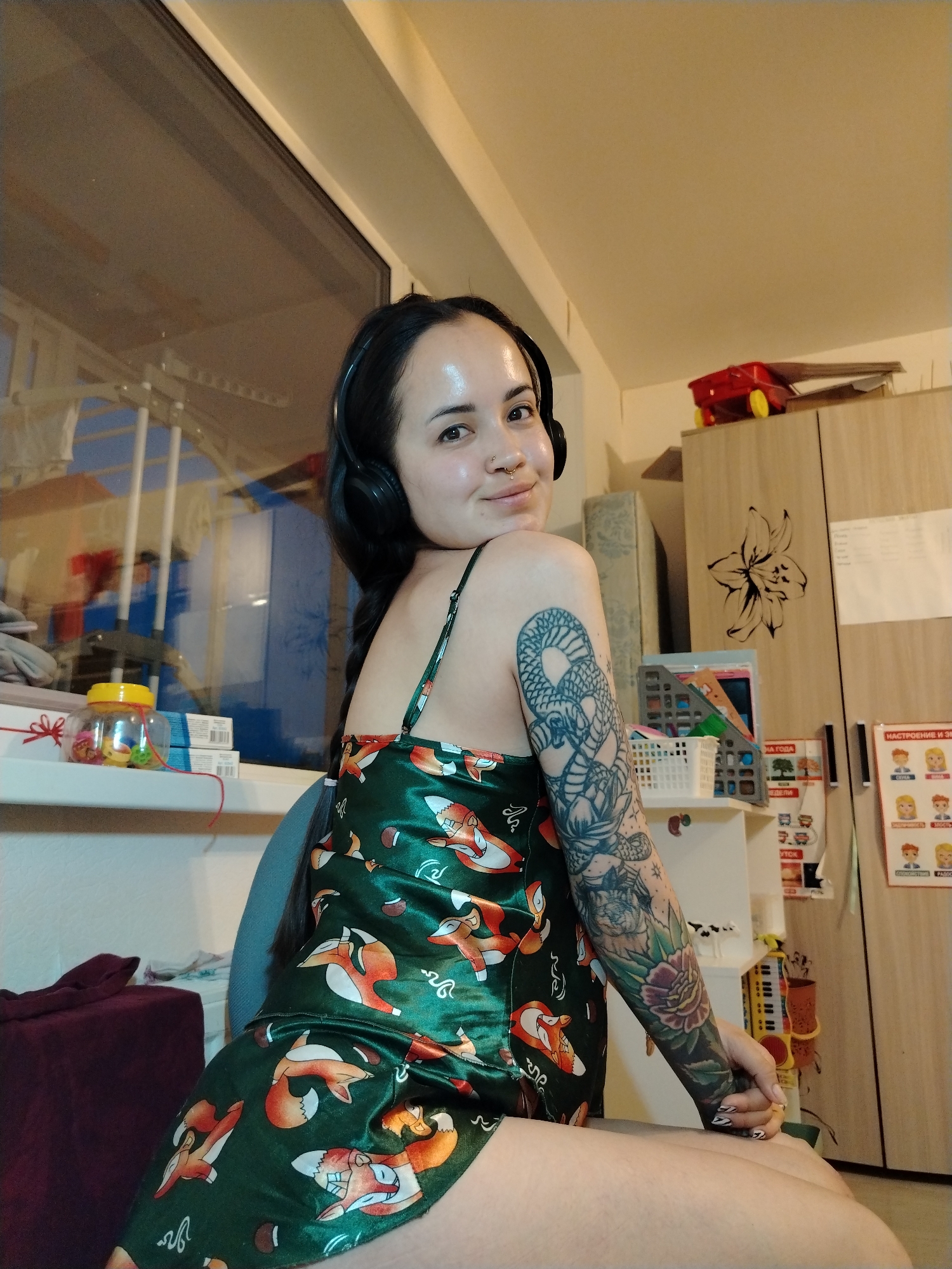 A woman with fair skin and dark hair is seated indoors. She is wearing a green pajama set patterned with orange fox illustrations, and has black headphones around her neck. Her left arm is heavily tattooed. The background includes a window showing an exterior scene with metal railings, and a beige cabinet with a flower decal. Educational posters in Russian are affixed to the cabinet. A red toy car is perched on top of the cabinet. Miscellaneous objects, including jars, are visible on a shelf near the window. The room is lit artificially. The scene appears to be in the city of Noviy, Russia based on the Russian language signage.