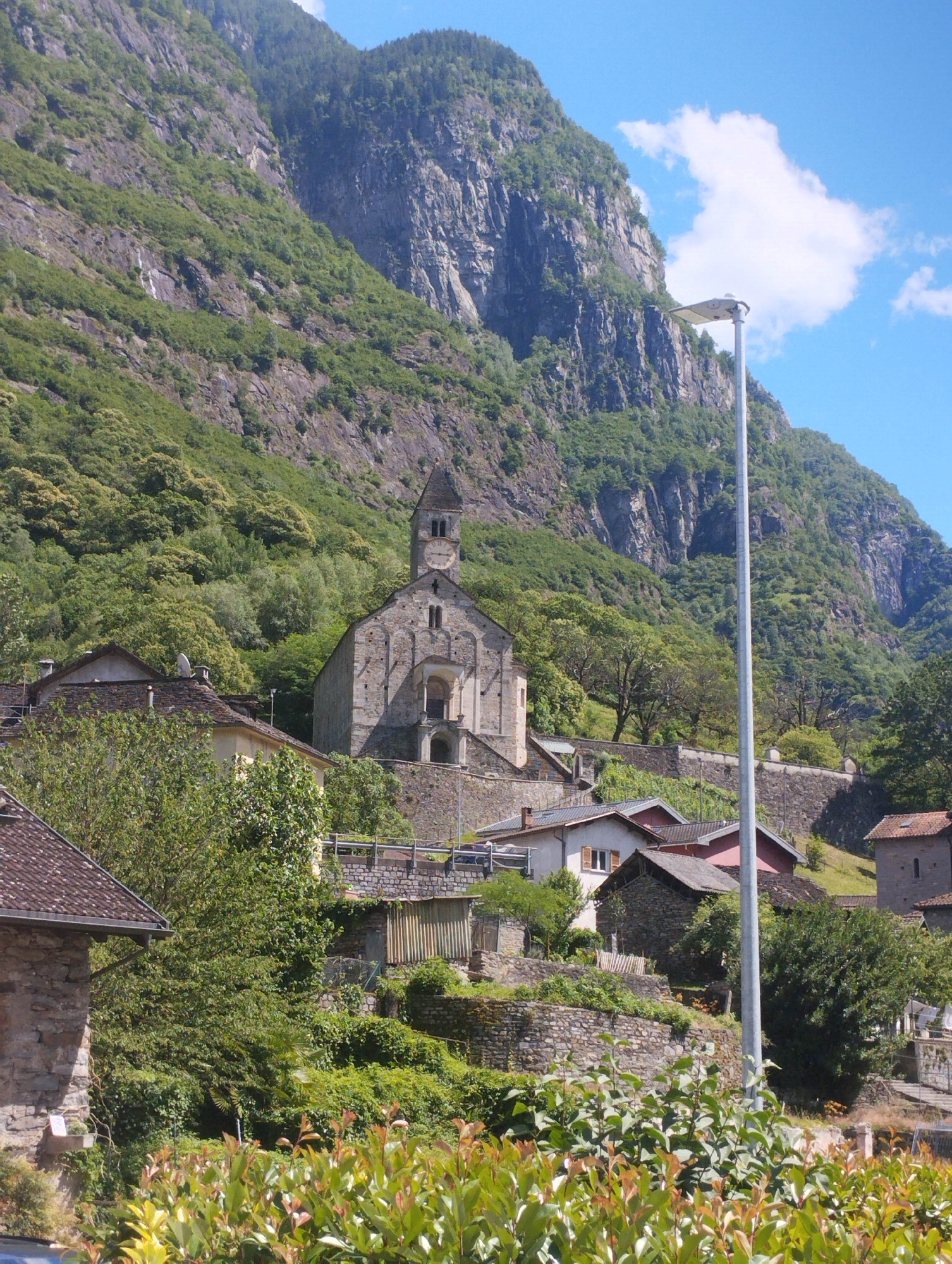 The image captures a picturesque mountain village under a bright, sunny sky with a few white clouds. In the midground, a prominent stone church with a tall, square bell tower and a clock face stands centrally, its architecture suggesting historical significance. The church is built into the hillside, surrounded by stone retaining walls and other traditional houses with tiled roofs.
The village is nestled on a steep, verdant mountainside, which dominates the background. The mountain features rugged, grey rock faces interspersed with abundant green trees and foliage, extending upwards into the distance. In the immediate foreground, a dense array of green and reddish-green bushes creates a vibrant border at the bottom of the frame.
A tall, modern grey lamppost stands conspicuously on the right side of the image, reaching high into the sky. Other houses are visible around the church, some partially obscured by trees and built on various levels of the slope, connected by stone walls and paths. The scene is bathed in bright daylight, casting clear shadows and indicating a sunny day, likely morning or early afternoon. No people or visible activities are depicted, giving the impression of a tranquil and scenic mountain settlement. No text is visible in the image.