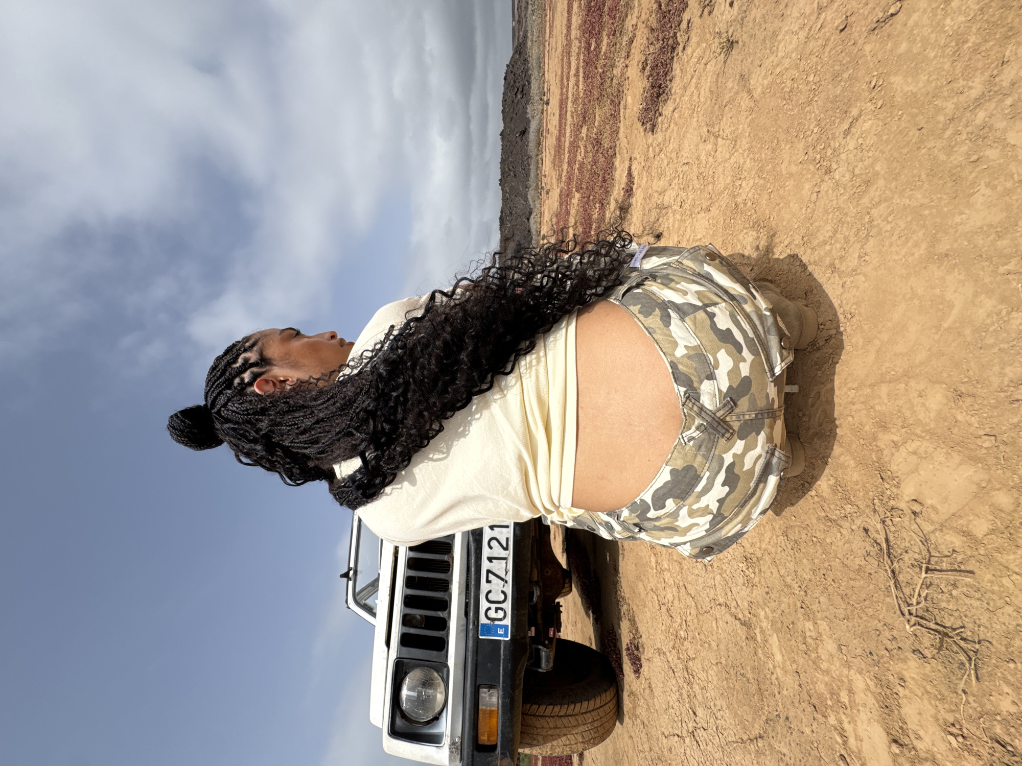 A person kneels on a vehicle's hood. The license plate number is GC7121. The environment appears to be an arid, outdoor location in Tinajo, Spain, with scrub vegetation and dirt ground visible. The sky is overcast with patches of blue. The person is wearing camouflage shorts and a light-colored top, with dark braided hair. The vehicle is white with a black bumper and grill.