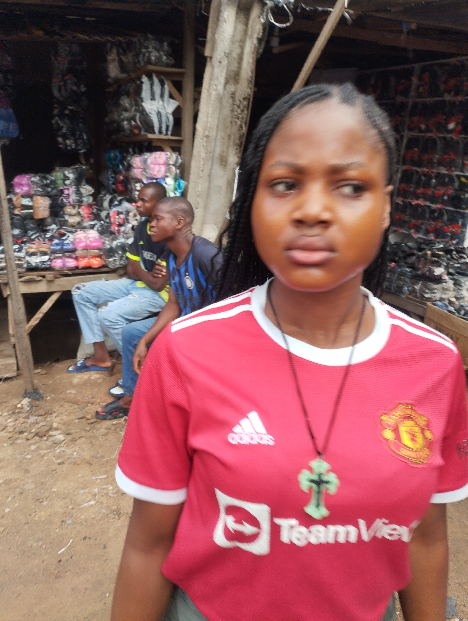 The image shows a young woman wearing a red Manchester United jersey with a white trim, standing in what appears to be a market area in Fegge, Nigeria. She is wearing a green cross necklace. Behind her, two men are sitting on a wooden platform, dressed in casual clothing. The platform appears to be part of a shop displaying various types of shoes or footwear. The background of the market area is filled with stacks of merchandise, suggesting a bustling commercial environment. The ground is dirt or sand. The lighting indicates it is daytime.
