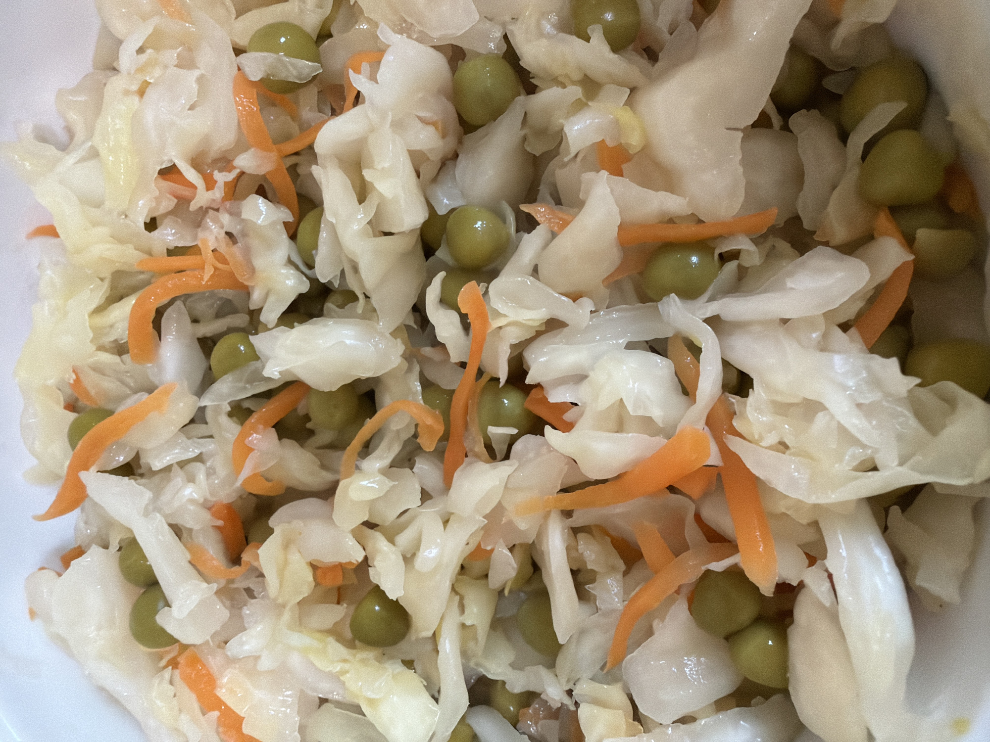 A close-up, overhead shot displays a serving of coleslaw. The salad is composed of finely shredded white cabbage, julienned carrots, and whole green peas. The ingredients appear to be mixed together in a white bowl, though the bowl itself is only partially visible at the edges of the frame. The lighting is even, and the focus is sharp, highlighting the texture and colors of the vegetables. The image is a still-life of food, with no people, animals, or discernible environmental context beyond the implied presence of a dishware. There is no information available in the image to suggest a location in Khabarovsk, Russia.