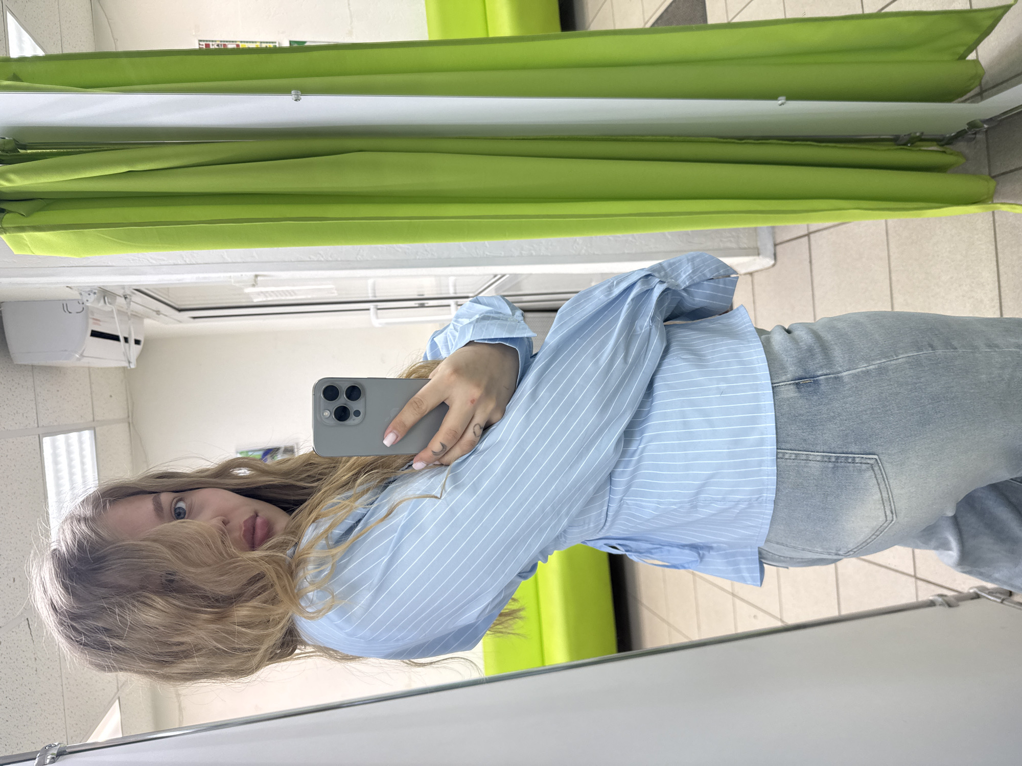 A woman with blonde hair is taking a selfie in what appears to be a dressing room or fitting room. She is wearing a light blue striped shirt and light blue jeans. She is holding a gray smartphone in her right hand. The background includes a green curtain or partition and a tiled floor. An air conditioning unit is visible on the wall. There is no information about the location being Chaikovsky, Russia.