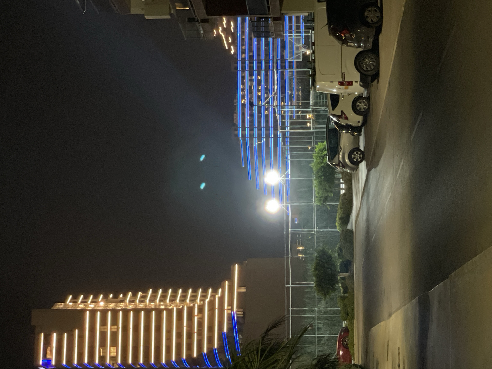 In the city of Trikomo, Northern Cyprus, an exterior night scene captures a street with several parked cars, including a white van and a dark SUV, on the right side of the asphalt road. Beyond the vehicles, a building is illuminated with blue horizontal light strips. Further to the left, a tall, tiered building is adorned with yellow horizontal light strips and blue accents along its edges. In the background, a large, transparent fence separates the road from a row of trees and bushes. The overall atmosphere is one of a modern urban environment at night, with architectural lighting creating a striking visual display.