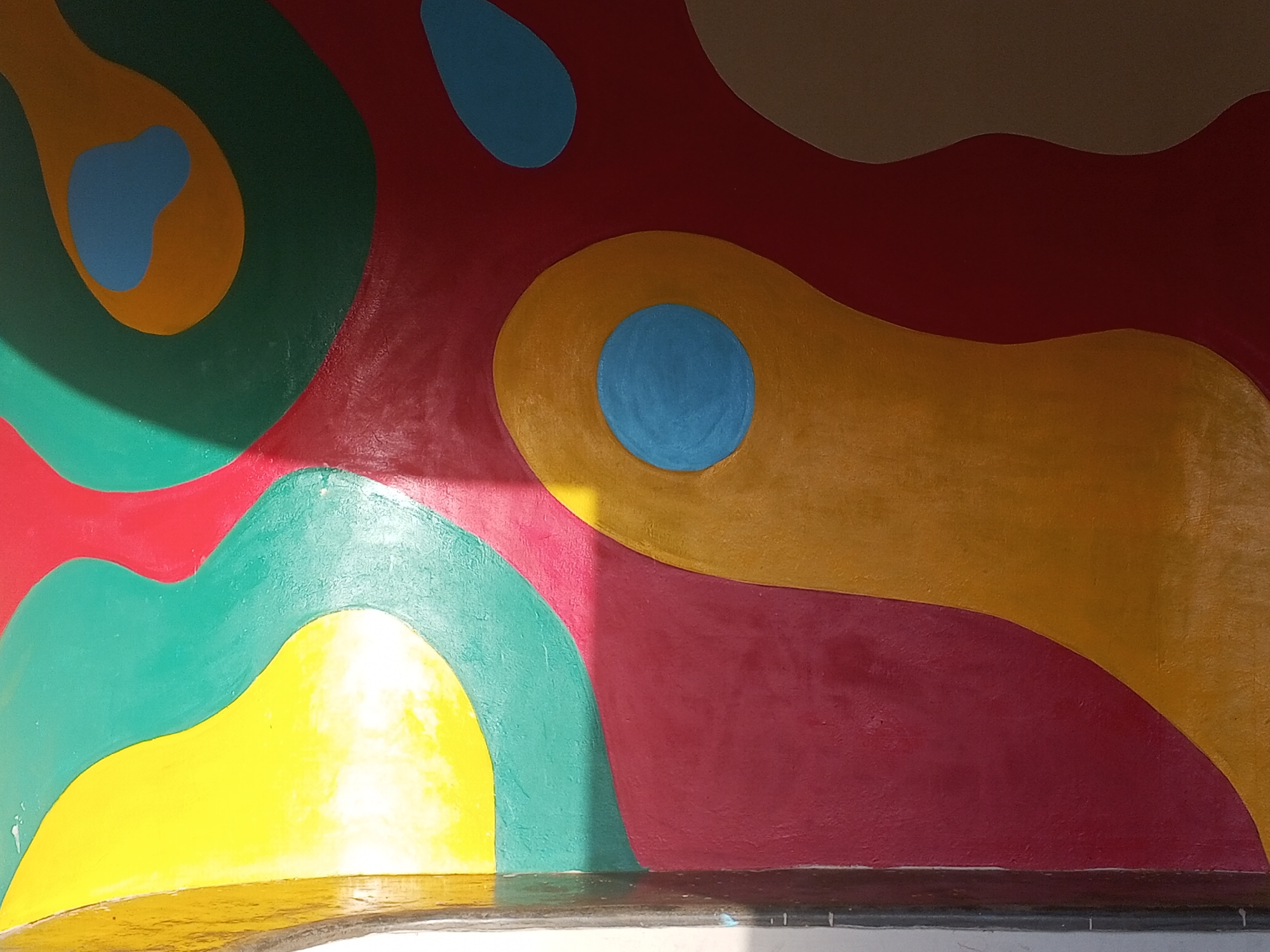 A brightly colored, abstract mural covers a wall in an unknown location. The mural consists of overlapping organic shapes painted in hues of red, yellow, green, and blue. Light casts shadows across the mural, adding depth. The bottom edge of the mural meets a wall with visible damage, but no people or other elements are visible. The location in Tawpo, Nigeria is not confirmed.