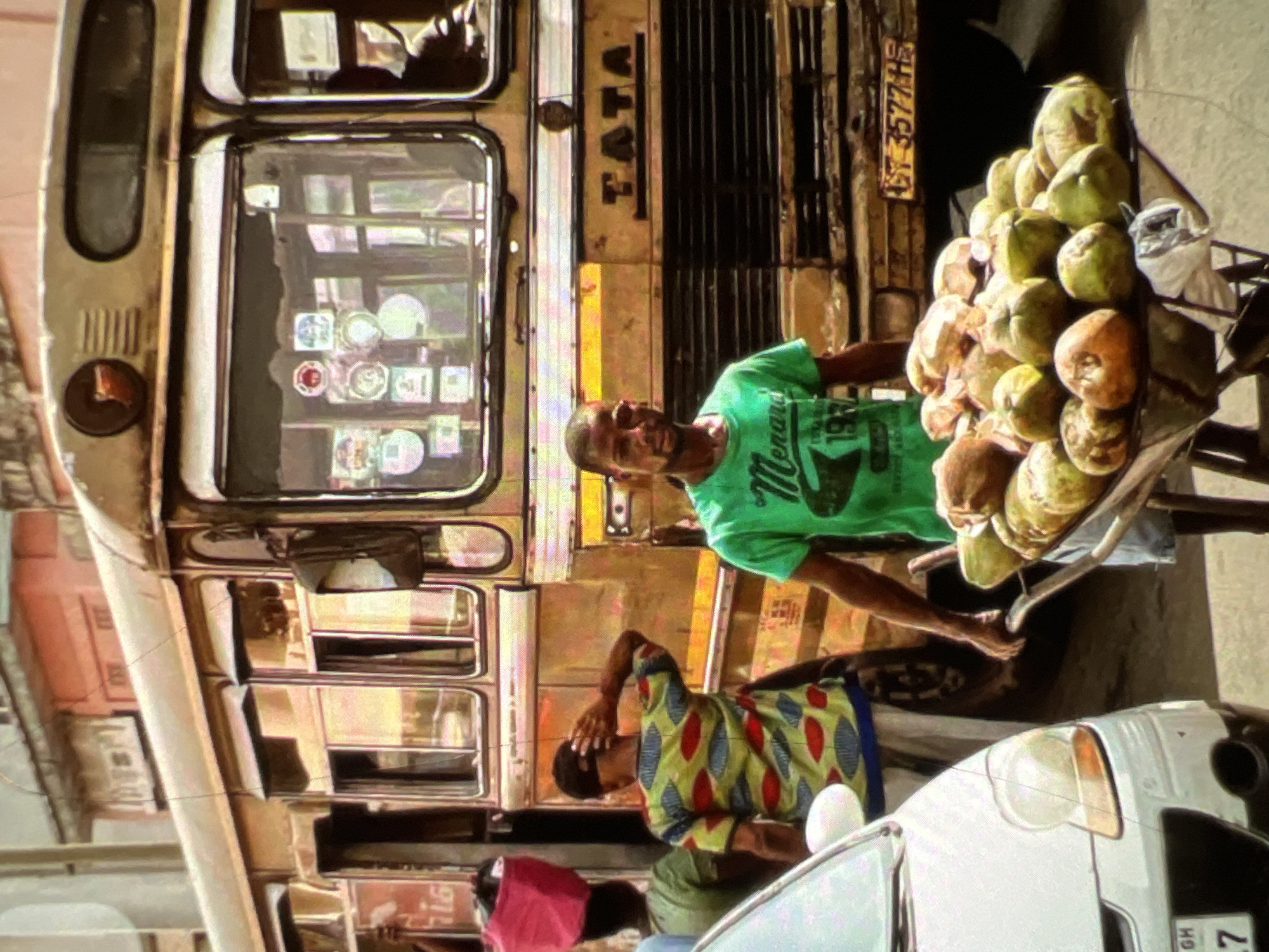 In the city of Lagos, Nigeria, a vendor pushes a wheelbarrow loaded with coconuts. The vendor, a dark-skinned male wearing a green t-shirt and shorts, is positioned on the right side of the image, facing forward. He is pushing the wheelbarrow towards the foreground. Behind him, a large, old yellow bus with the brand name "TATA" visible on its side is parked. The bus has several windows, some of which reflect the surroundings. To the left of the bus, another person is visible in a colorful patterned shirt, with their back to the camera. A white car is parked on the far left of the frame. The scene appears to be a street in a busy urban environment.