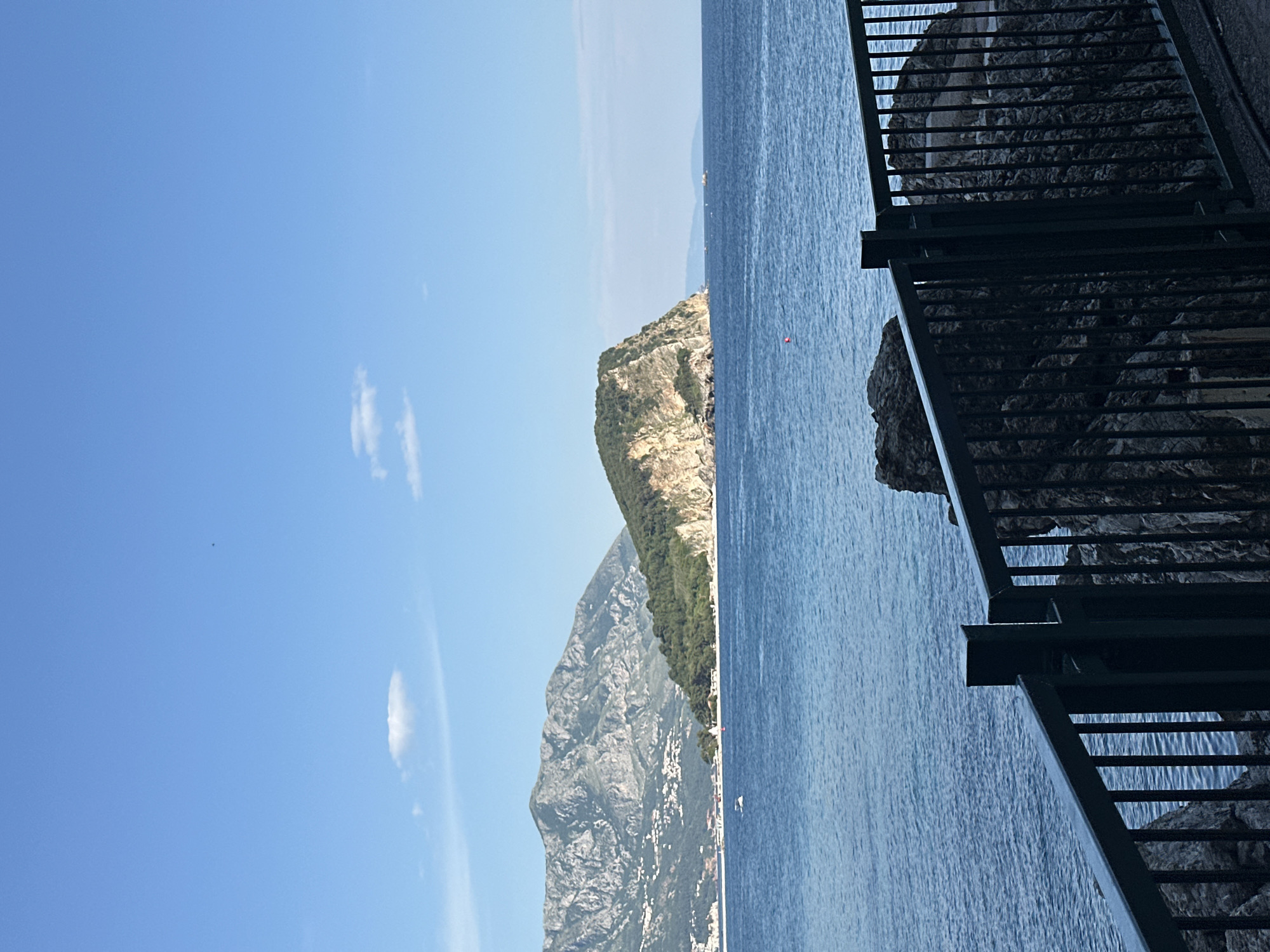 The image depicts a coastal scene, possibly near Budva, Montenegro, given the rugged terrain and blue water. To the right, a dark railing suggests a viewpoint or walkway. Below the railing, rough rock formations are visible. The sea is a deep blue, stretching out to the horizon. In the mid-ground, a cliff covered with vegetation rises from the water. Further inland, a larger mountain range is visible, dotted with what appears to be white buildings. The sky is a clear blue with a few scattered clouds. A small, red object is visible in the water near the cliff. The image is rotated 90 degrees counter-clockwise.