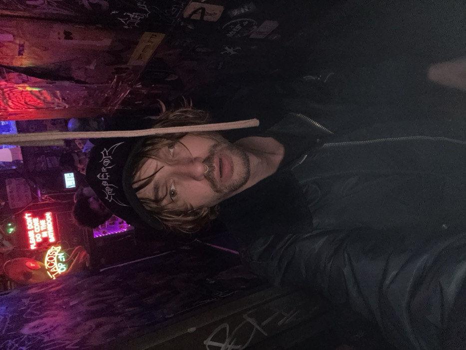 A man with shoulder-length blonde hair, a nose ring, and a goatee is wearing a black jacket and beanie. He is holding a thin, light-colored stick in front of his face. Behind him, there's a dimly lit space resembling a bar or venue. A neon sign says "PLEASE DON'T DO COKE IN THE BATHROOM". A second sign says "TACOS OPEN". The scene has graffiti and stickers on the ceiling and walls, creating a cluttered and edgy atmosphere.