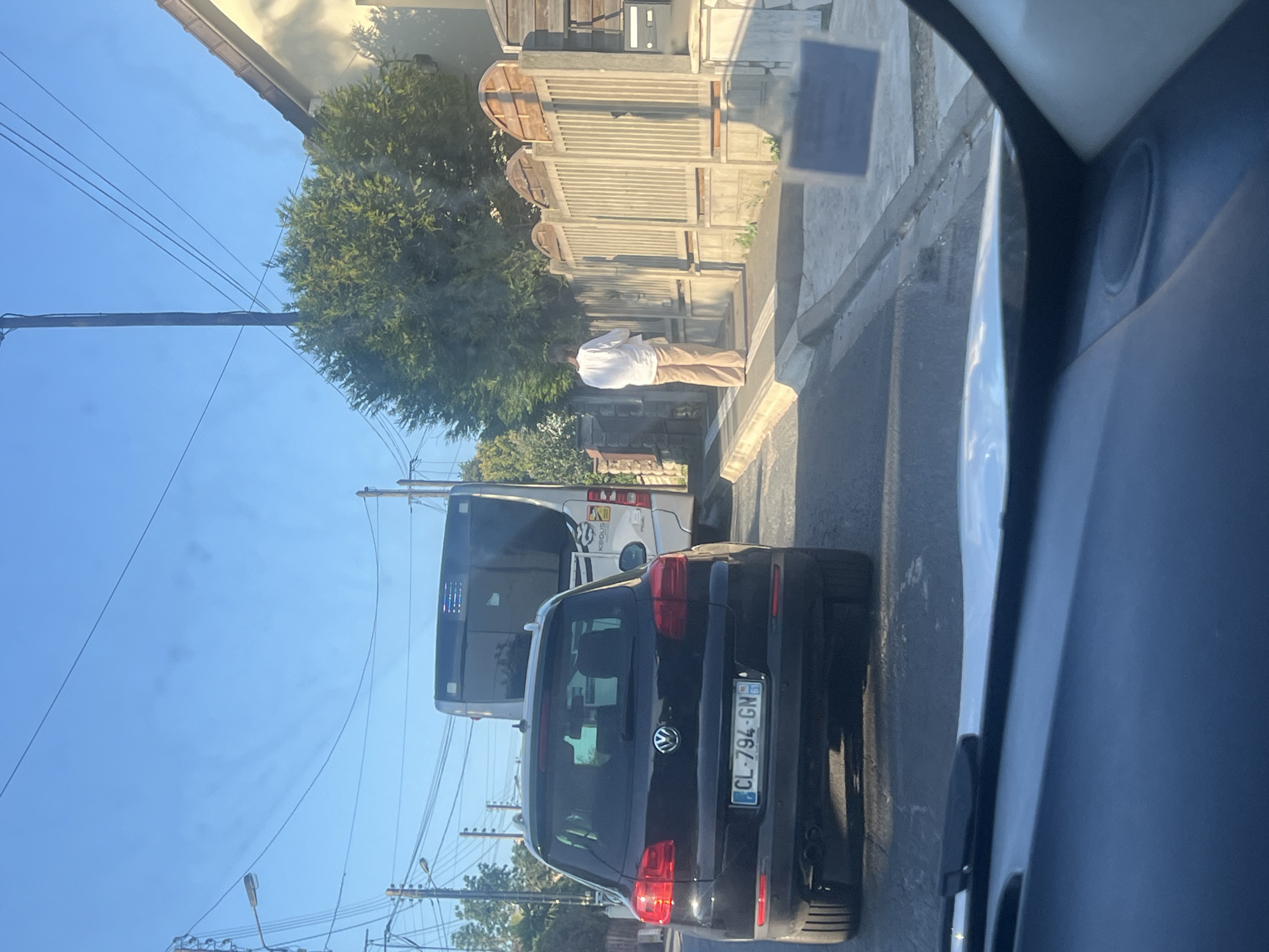 The image captures a street scene in Gagny, France, likely on a sunny day given the clear blue sky. In the foreground, a black Volkswagen Tiguan is parked, its rear visible, displaying a French license plate: CL-794-GN. Behind it, a white bus, possibly a public transport vehicle, is also stopped. To the right of the bus, a person dressed in a white long-sleeved shirt and beige pants stands on a sidewalk next to a fence. The fence appears to be made of concrete and wood, with arched wooden panels at the top. The street is paved, and a sidewalk runs along the right side of the image. Overhead, power lines and a streetlight are visible against the sky. The overall impression is that of a quiet suburban street during daylight hours.