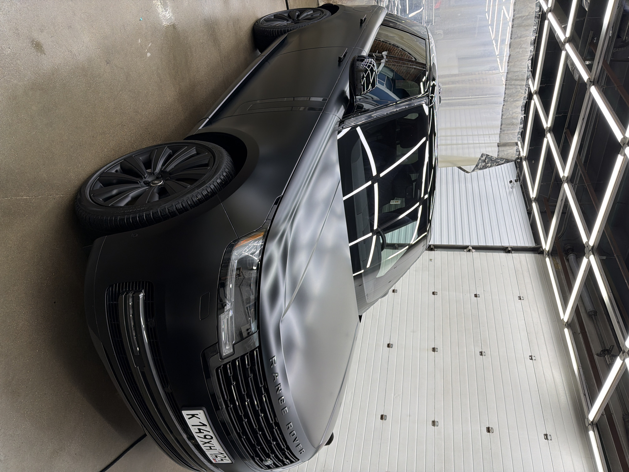 The media file presents a black matte Range Rover SUV parked indoors, likely within a car wash or detailing facility in Krasnoyarsk, Russia. The vehicle is positioned with its front facing the viewer's right, showcasing its driver's side and front quarter panel.
The Range Rover features a sleek matte black body finish, complemented by glossy black accents on its grille, headlight surrounds, and window trim. It is fitted with matching black, multi-spoke alloy wheels. The word "RANGE ROVER" is visibly embossed in black letters on the front of the hood. A Russian license plate, "K149XH 124 RUS," is attached to the front bumper, with "124" indicating the Krasnoyarsk Krai region.
The setting is characterized by bright, artificial overhead lighting arranged in a prominent grid pattern. These lights produce striking, geometric reflections on the car's windows, side panels, and the subtly textured matte hood, highlighting the car's contours. The floor appears to be concrete, showing some signs of dampness, consistent with a car cleaning environment. The background consists of white, possibly corrugated or paneled walls or a garage door.
The car is stationary, suggesting it is either undergoing or has recently completed a cleaning or detailing process. No people are visible in the image. The bright indoor illumination precludes any specific indication of the time of day or external weather conditions.