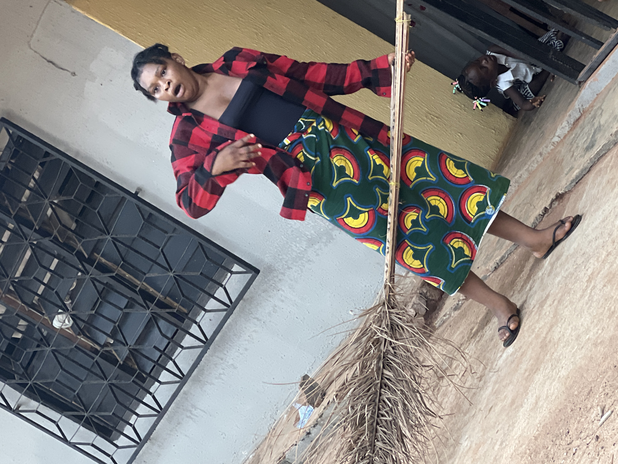 An outdoor, slightly tilted full-shot captures a woman and a child in what appears to be a residential or public space in Onitsha, Nigeria. The central figure is a woman with braided hair, dressed in a red and black plaid shirt worn open over a black tank top, and a vibrant green skirt adorned with yellow and red circular patterns, indicative of African print fabric. She wears black flip-flops. Her mouth is open in an expression that suggests surprise, speech, or animated conversation. She holds a tall, thin pole-like object, which expands at the bottom into a dense cluster of dried, light brown palm fronds, resembling a traditional broom or sweeping tool.
In the upper right background, a small child with braided hair, adorned with colorful ties, is seen peeking out from a dark, elevated opening, possibly a window or doorway. The child wears a white top and appears to be observing the scene.
The setting includes a light grey wall on the left, featuring a black metal security grate with a geometric pattern. Above the grey wall, and behind the woman, is a section of yellow-ochre colored wall. The ground beneath the woman's feet is a reddish-brown, uneven dirt or concrete surface. The scene is illuminated by daylight. There is no visible text.