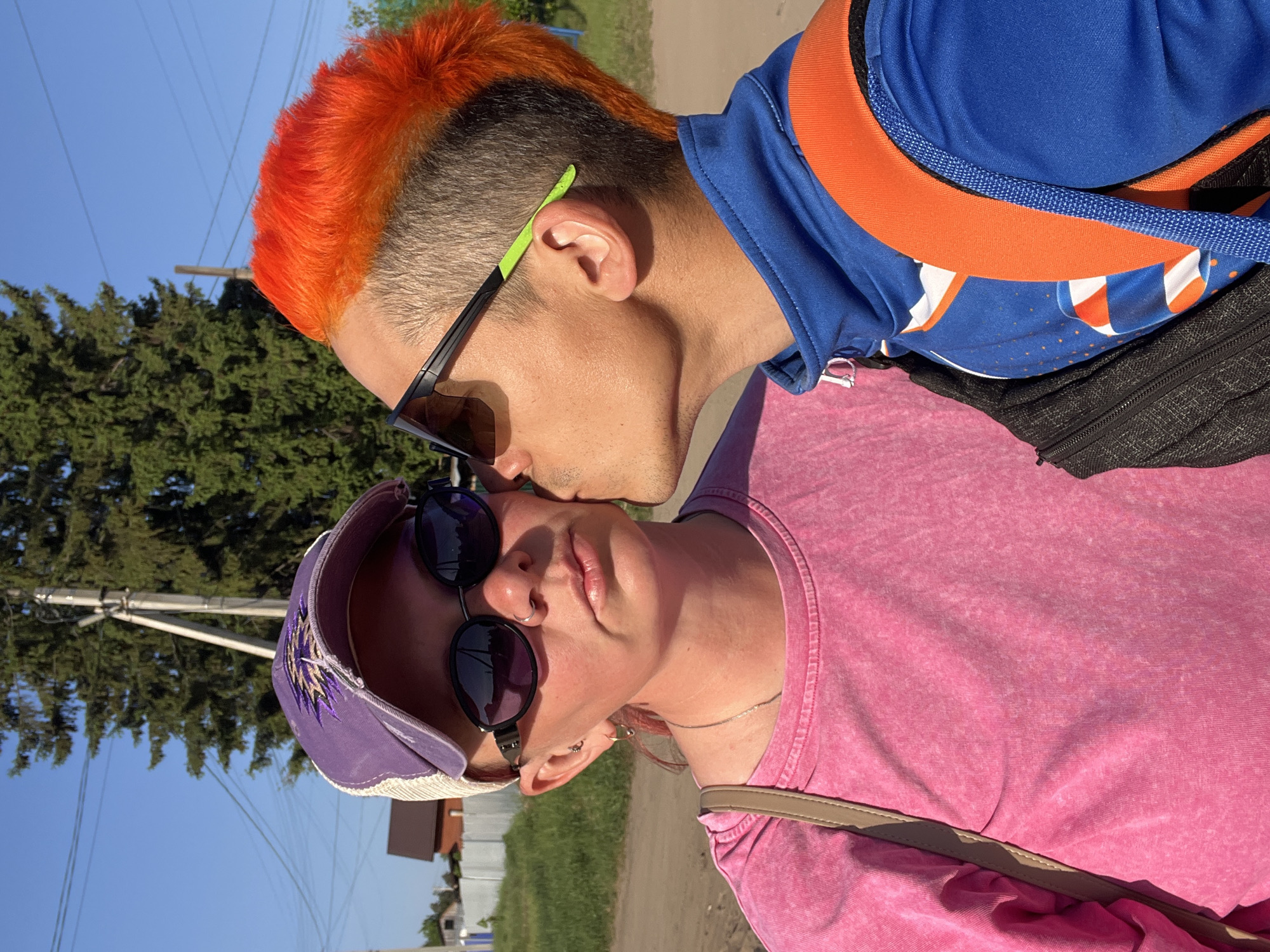 A close-up shot captures two individuals in an outdoor setting. The person on the right, with a bright orange Mohawk hairstyle, wears sunglasses with green and black frames and a blue and orange jacket with a black backpack. They are leaning in to kiss the forehead of the person on the left.
The person on the left wears a pink t-shirt, a purple baseball cap with a white mesh back, and sunglasses. A tan strap of a bag is visible across their shoulder. They have piercings in their nose and ear.
The location appears to be near a road with buildings and trees in the background. Based on prior knowledge, it is highly probable that this photo was taken in the city of Chaikovsky, Russia, a mid-sized industrial city located in the Perm Krai region of Russia. The presence of a visible power line and a partially visible residential building support this localization.