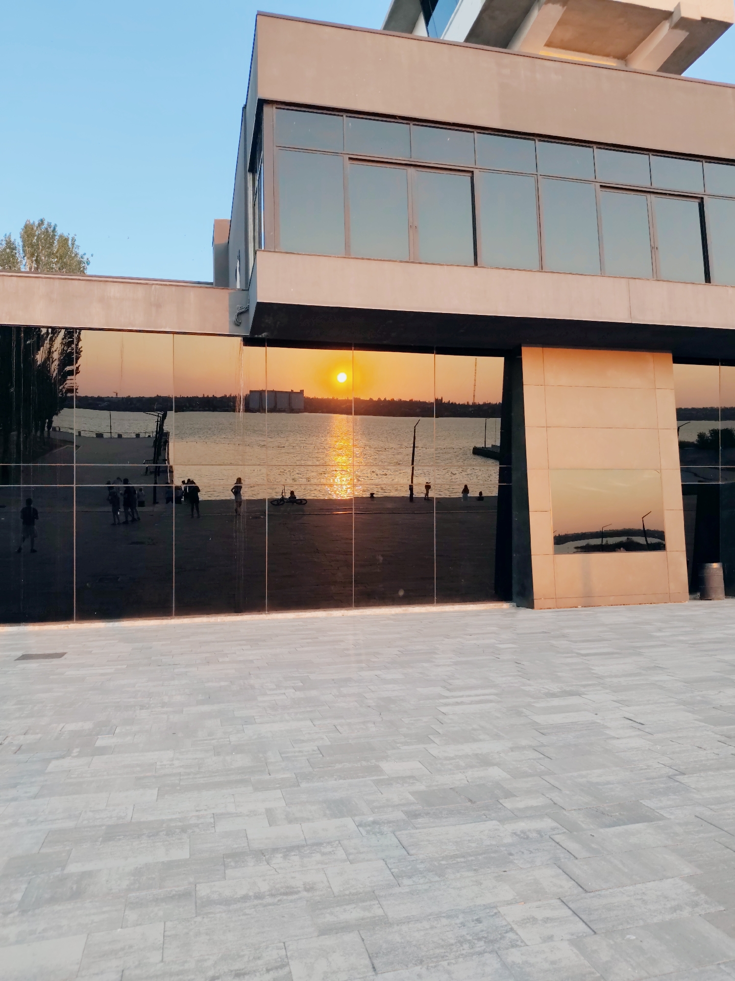 The image depicts the facade of a modern building with large, reflective glass windows, likely situated along a waterfront in Mykolaiv, Ukraine. The glass reflects a vibrant sunset over a body of water, with the sun appearing as a bright orange orb. Silhouettes of people are visible in the reflection, some standing and others appearing to be walking. A bicycle is also visible in the reflection. The building itself has a minimalist design with smooth, beige-colored exterior panels. The foreground is a paved walkway made of light-colored rectangular stones. The sky above is a clear, pale blue, with a few faint birds visible. The overall scene conveys a sense of a serene evening at a public space by the water.