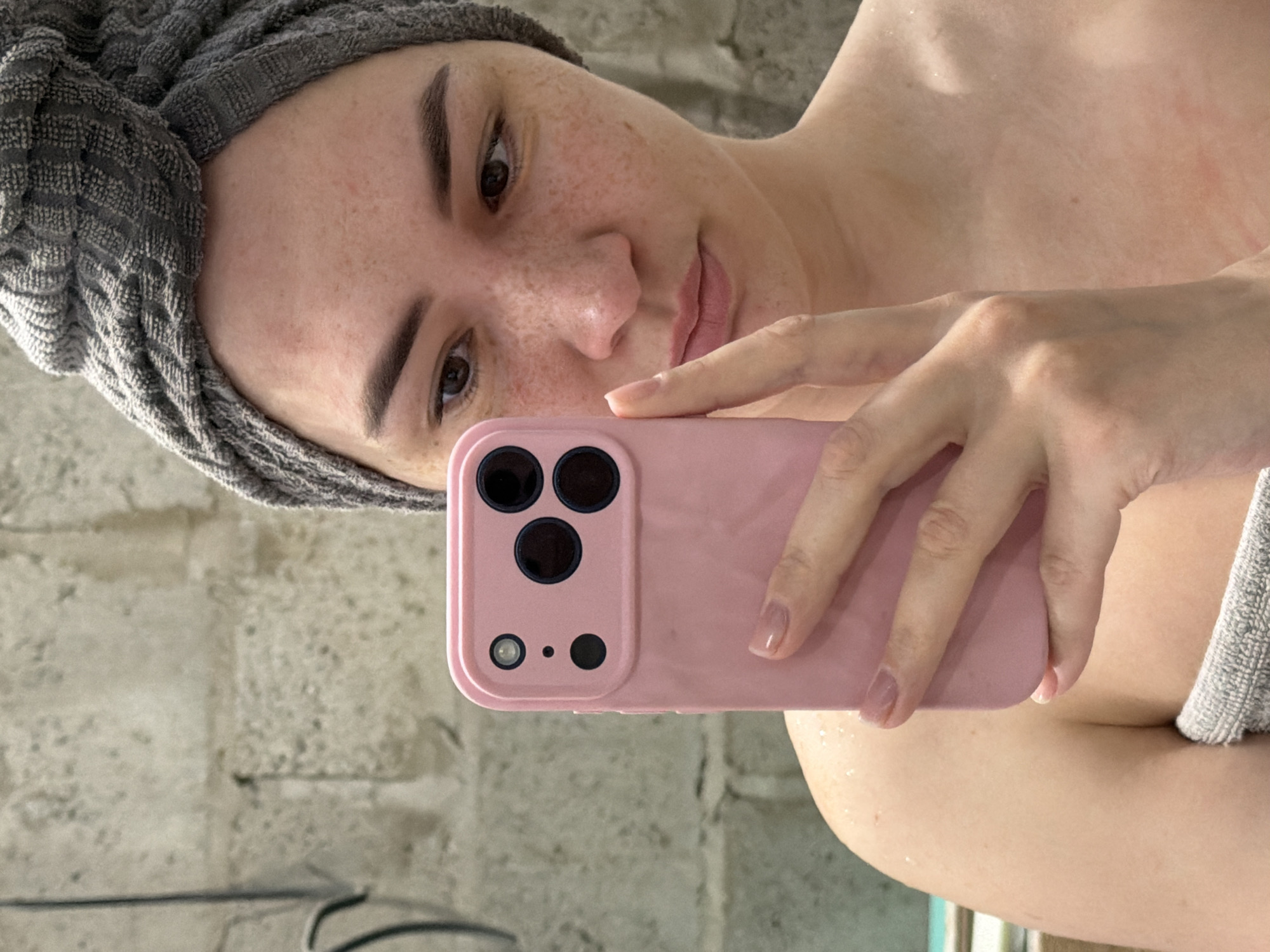 A woman with freckles and a grey towel wrapped around her head is holding a pink cell phone up in front of her face, presumably taking a selfie. The background appears to be a concrete wall. The exact location in Chaikovsky, Russia is indeterminable from the image.
