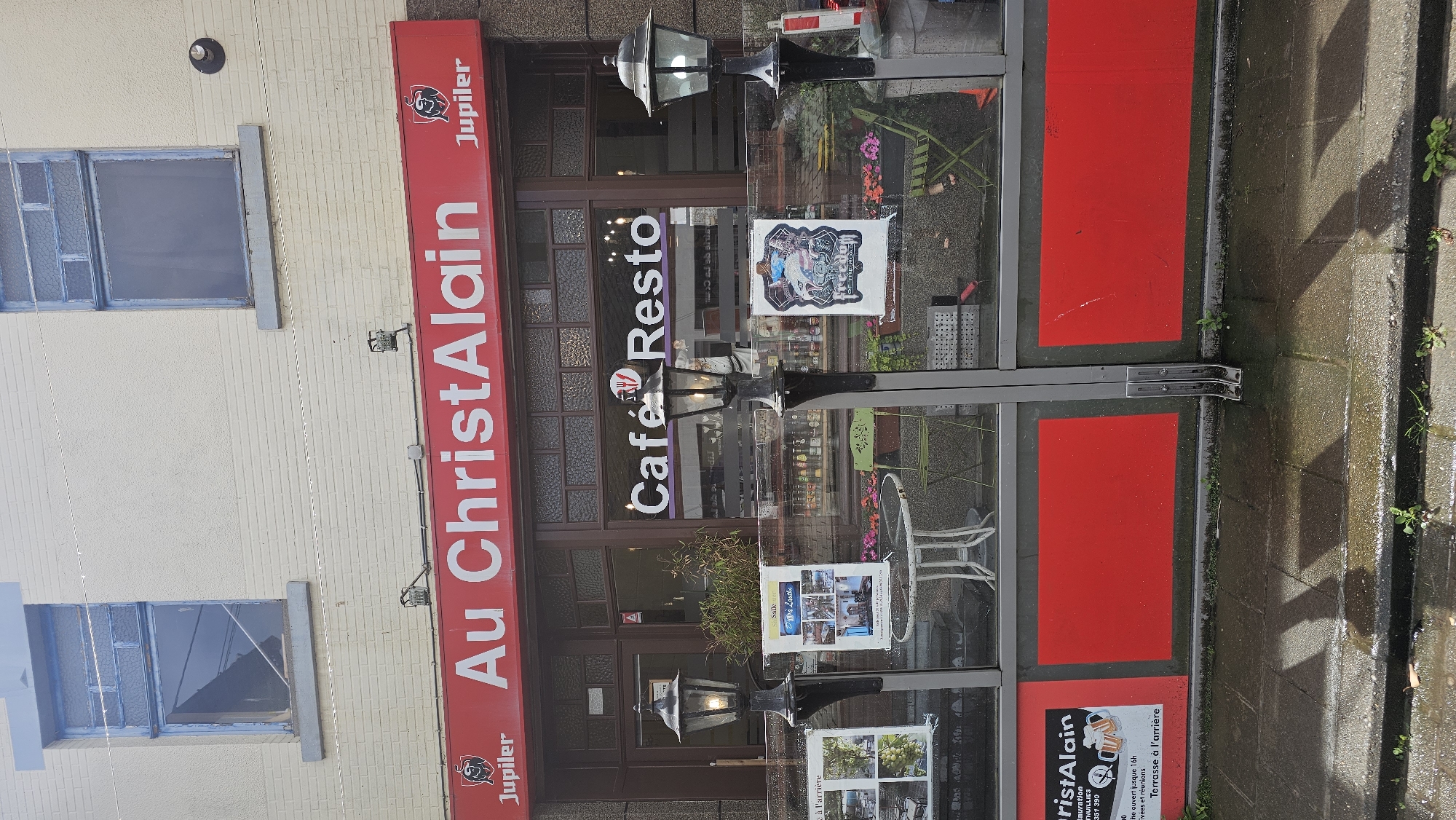 This media file captures the exterior of a cafe or restaurant named "Au ChristAlain" in Thuin, Belgium. The scene is viewed from across a wet pavement, suggesting recent rain or damp conditions.
The establishment features a prominent red banner sign along its facade with "Au ChristAlain" written in white lettering, accompanied by the Jupiler beer logo. Below this, the entrance area consists of large glass windows and a dark-framed door. Inside the windows, another sign reads "Café Resto" in white and purple. Various posters are displayed on the glass, one with the text "Passez à l'arrière" (Go to the back) and "Terrasse à l'arrière" (Terrace at the back), indicating additional seating. Other posters show images of the interior or dishes offered, and one clearly mentions "Thuin" as part of an address or contact information.
Outdoor seating is visible through the glass, including small tables and chairs, some green and white. Two decorative lampposts are positioned outside the entrance. The building itself is light-colored, possibly painted brick or stucco, with two blue-framed windows on the upper floor. The pavement directly in front of the establishment features red rectangular sections integrated into the ground, possibly part of an outdoor dining area or a decorative element. Small weeds are growing in cracks in the concrete pavement. The time of day appears to be daylight, but without direct sunlight.
Visible text includes:
- "Au ChristAlain"
- "Jupiler" (logo and text)
- "Café Resto"
- "Passez à l'arrière"
- "Terrasse à l'arrière"
- "Rauration THUILLIES"
- "Tel: 071 591 390"
- "Salle ouvert jusque 16h" (Room open until 4 PM)
- "Réunions et arrivées" (Meetings and arrivals)
- "La Lutèce" (on a poster)
- "10h" (possibly an opening time)