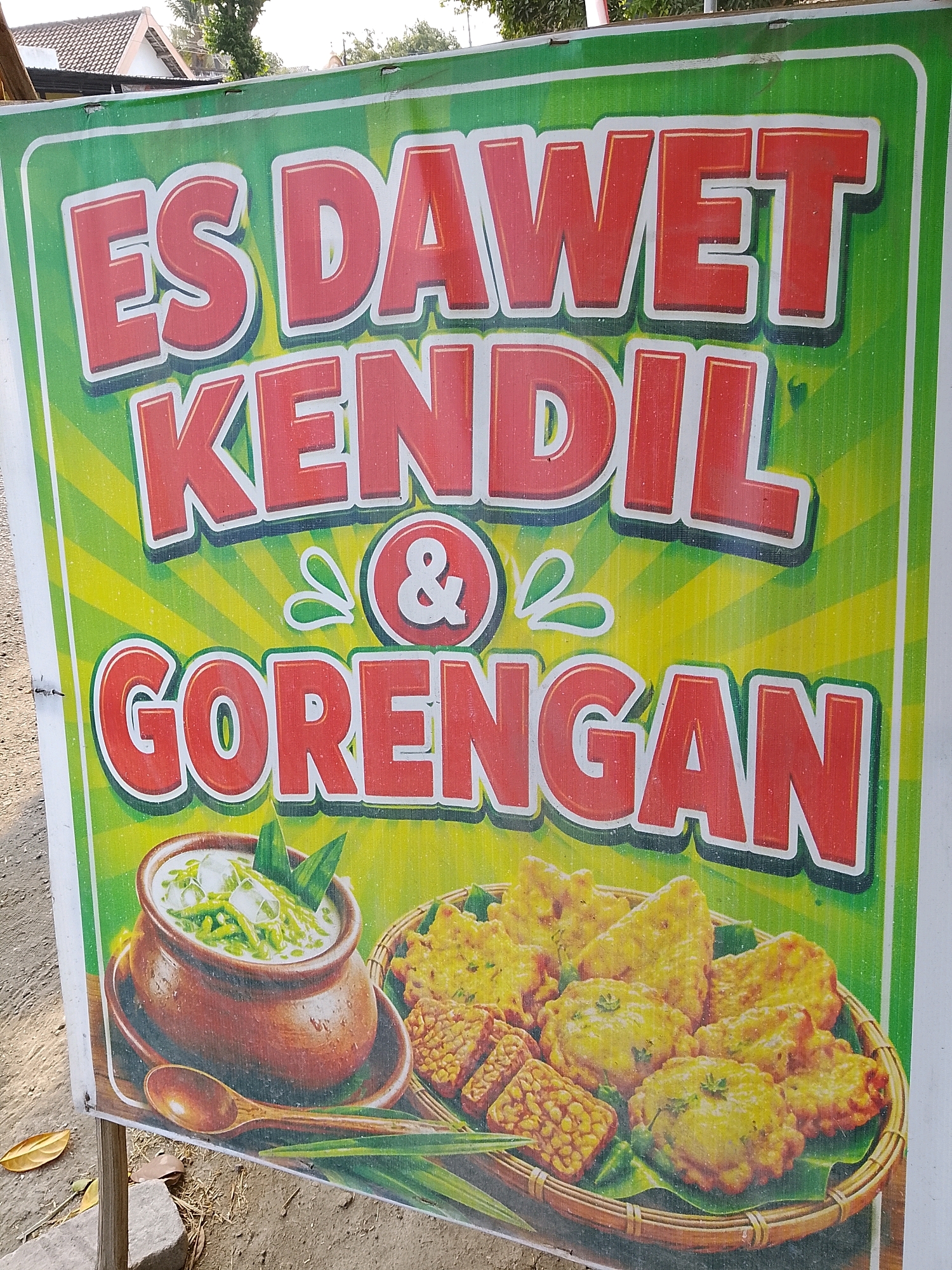 This is a photograph of a banner advertising "ES DAWET KENDIL & GORENGAN" in Banaran, Indonesia. The banner features large, red, stylized text against a vibrant green and yellow striped background with sunburst rays. The text reads "ES DAWET KENDIL" in the top two lines, followed by an ampersand symbol "&" with decorative green flourishes, and then "GORENGAN" on the bottom line. Below the text, to the left, is an illustration of a clay pot filled with a creamy white liquid, green jelly-like strands, and ice cubes, garnished with pandan leaves. A wooden spoon rests in front of the pot. To the right of the pot is an illustration of a woven basket filled with various fried snacks, including what appear to be tempeh and other fritters, also placed on banana leaves. The banner is attached to wooden posts and is outdoors, with a partially visible roof and foliage in the background. The ground in front of the banner is composed of dirt, gravel, and dry leaves.