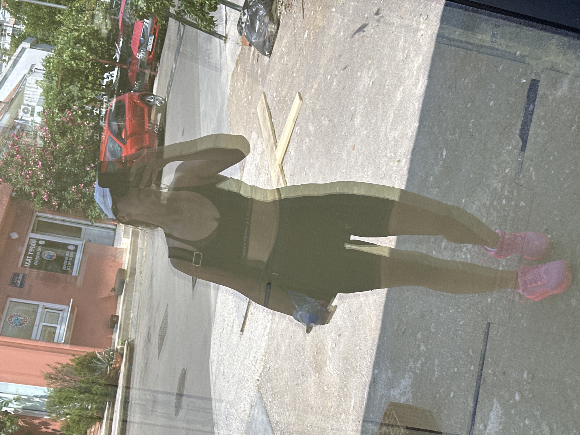 A woman in athletic wear, including a black sports bra and black biker shorts, is reflected in a glass surface. She is wearing pink athletic shoes and a light-colored cap. In her right hand, she holds a plastic-wrapped bottle of water. Behind her, the reflection shows a street scene with several red cars parked. One car is a bright red hatchback, and another appears to be a sedan. There is a building with signs, including one that reads "HAPPY TAXI" in an advertisement. The ground is paved, and there are some potted plants and trees visible. In the foreground of the reflection, two wooden planks are laid on the ground, forming a cross shape. A black garbage bag is also visible in the background of the reflection. The overall scene appears to be in an urban environment. The image has a slightly distorted and overlaid effect due to it being a reflection. The presence of "HAPPY TAXI" suggests a location where taxi services are available, and given the architecture and vegetation, it is consistent with a Mediterranean or Balkan setting, such as Budva, Montenegro.