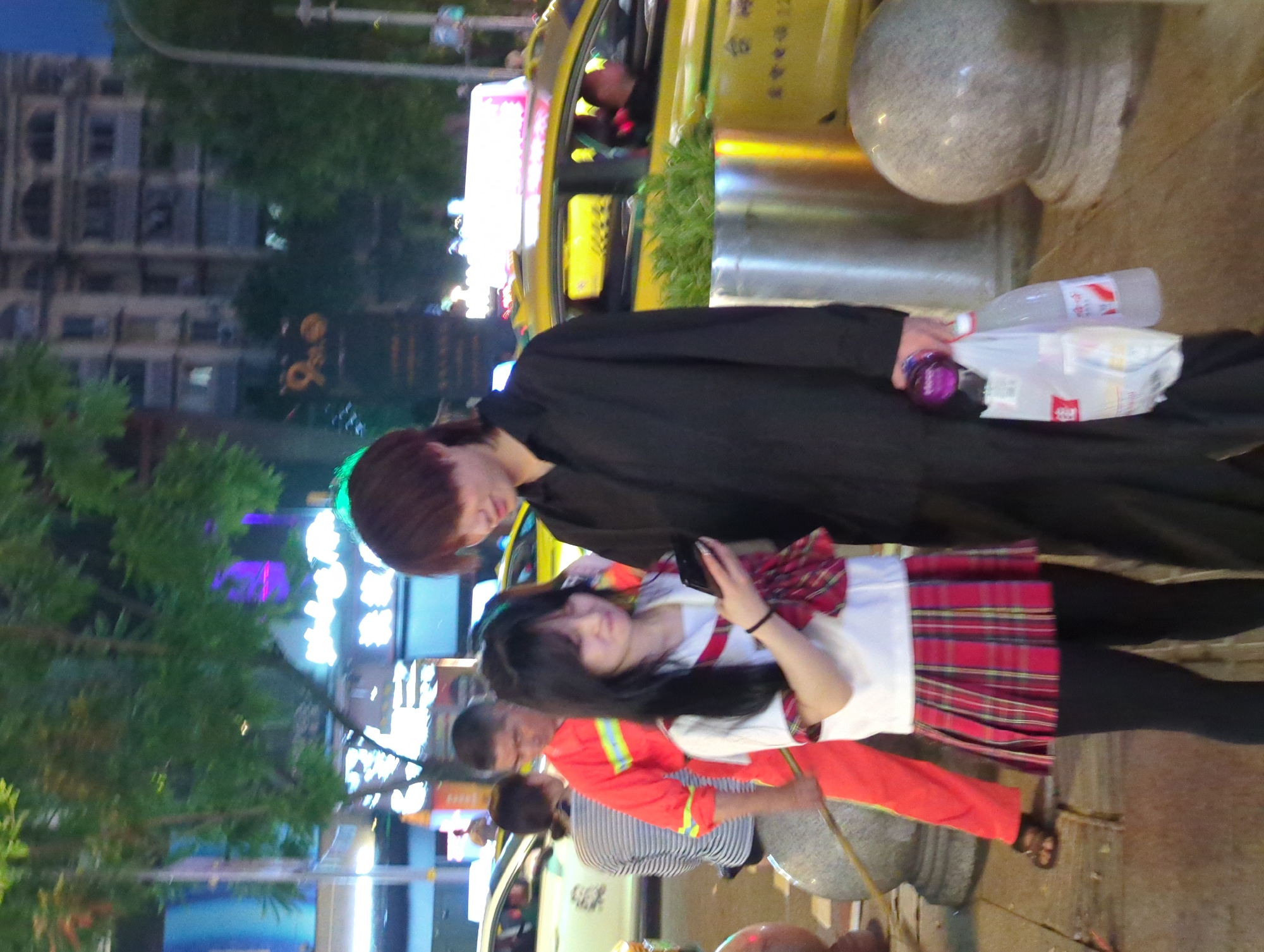 At night, in the city of Longta, China, a young woman in a red plaid skirt and black leggings is looking at her phone. She is standing next to a man in a black shirt and pants. In the background, there are several cars, including a yellow taxi, and buildings with illuminated signs. A person in an orange uniform is visible behind the young woman. The environment appears to be a street or public area, with trees and streetlights.
