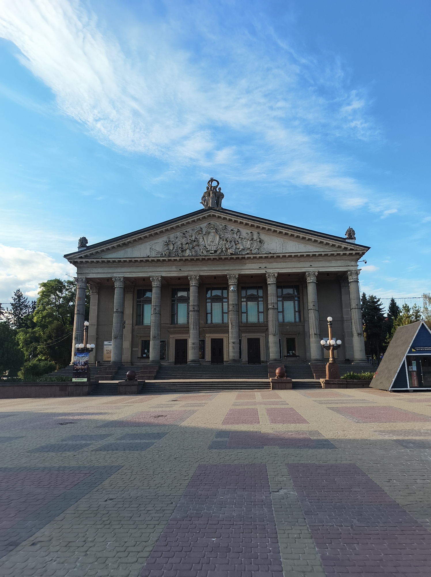 The image shows the Ternopil Regional Philharmonic building in Ternopil, Ukraine. The building features a grand neoclassical facade with a portico of Corinthian columns and a pediment adorned with sculptures. A paved plaza with a patterned surface is in the foreground, and a small triangular tourist information booth is visible on the right side of the plaza. The sky is blue with scattered clouds, and trees are visible behind the building. The scene is captured from a low angle, emphasizing the size and grandeur of the philharmonic building. No people are visible in the image.