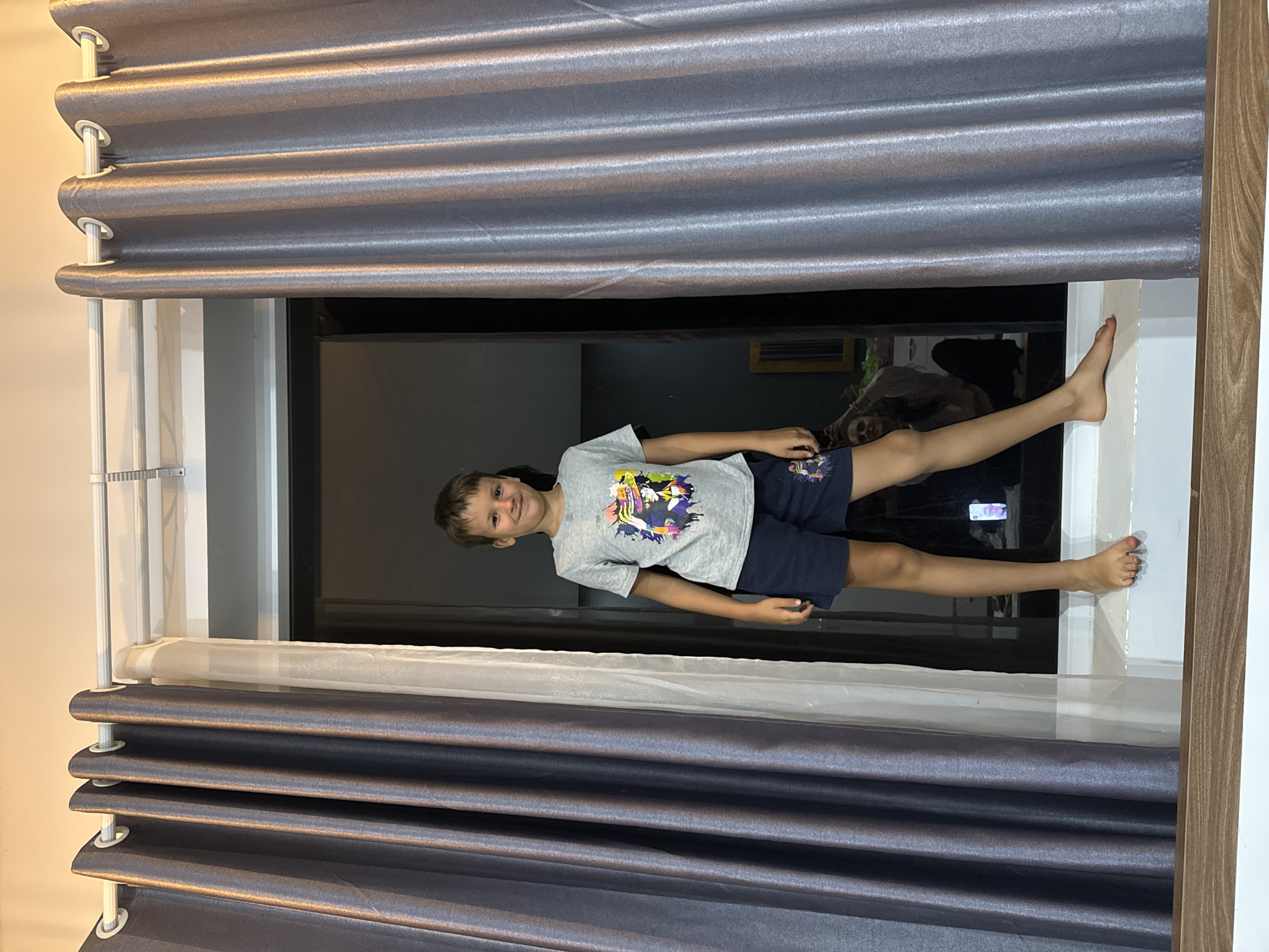 A child lies across a windowsill in Da Lat, Vietnam. The child, wearing a grey t-shirt and navy shorts, is positioned horizontally in the window frame. Grey, pleated curtains hang above and below the window. Outside the window is a blurred, dark background. The window frame is dark with a white painted sill and a wooden frame.