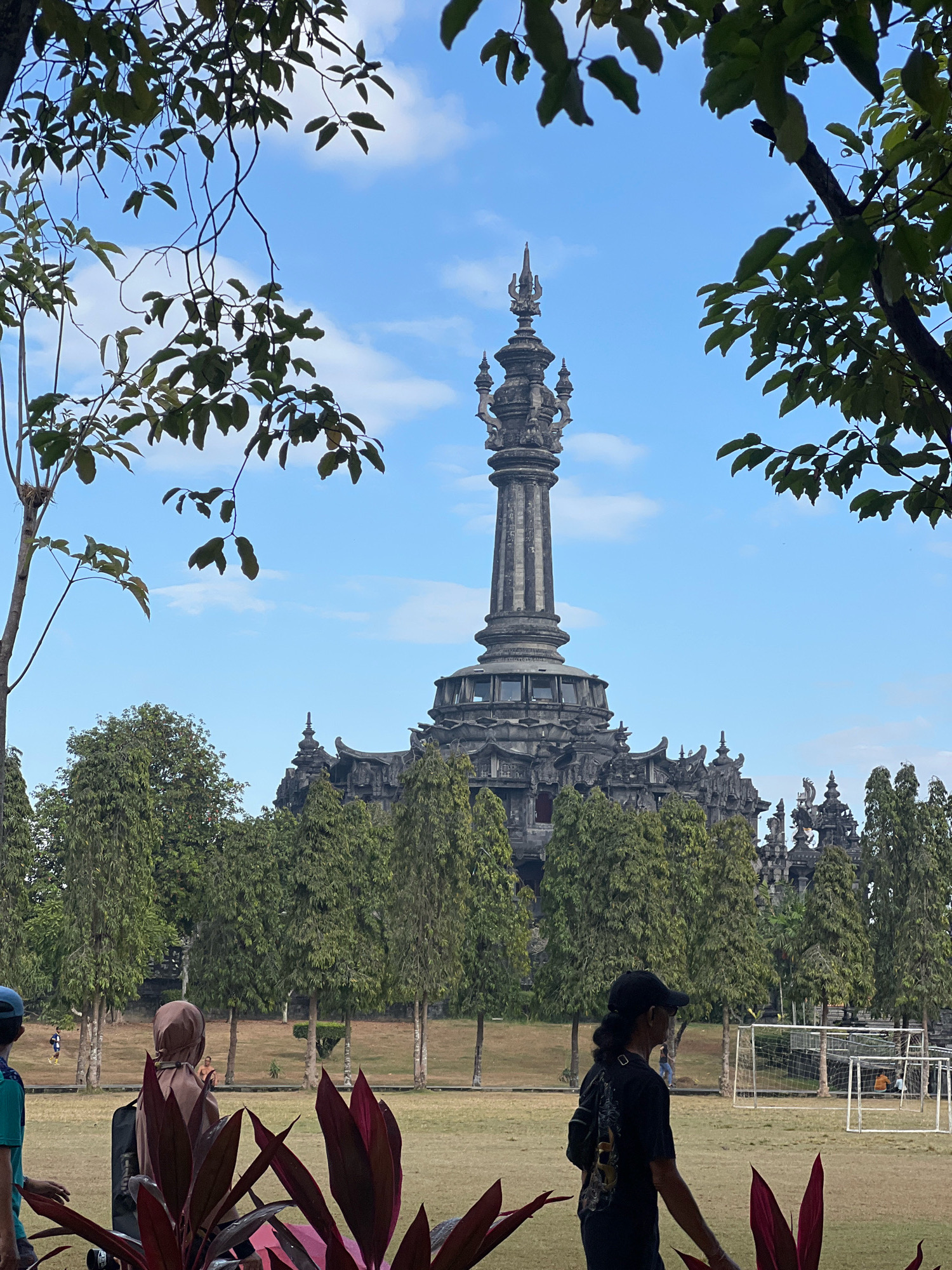 The image shows a monument in Denpasar, Indonesia, possibly the Bajra Sandhi Monument, set against a backdrop of trees and a blue sky with scattered clouds. In the foreground, lush green foliage partially obscures the view. Several people are visible in the scene. To the left, a person wearing a blue cap and a teal shirt is partially visible. In the middle ground, a person wearing a brown hijab stands facing away from the camera. To the right, a person wearing a black t-shirt and a black cap walks away from the camera, carrying a black bag slung over their shoulder. Behind them, a soccer field with white goalposts is visible. The monument itself is a tall, ornate structure with multiple levels and intricate carvings, characteristic of Balinese architecture. The overall scene depicts a public space with people enjoying the outdoors, with the monument as a central focal point.