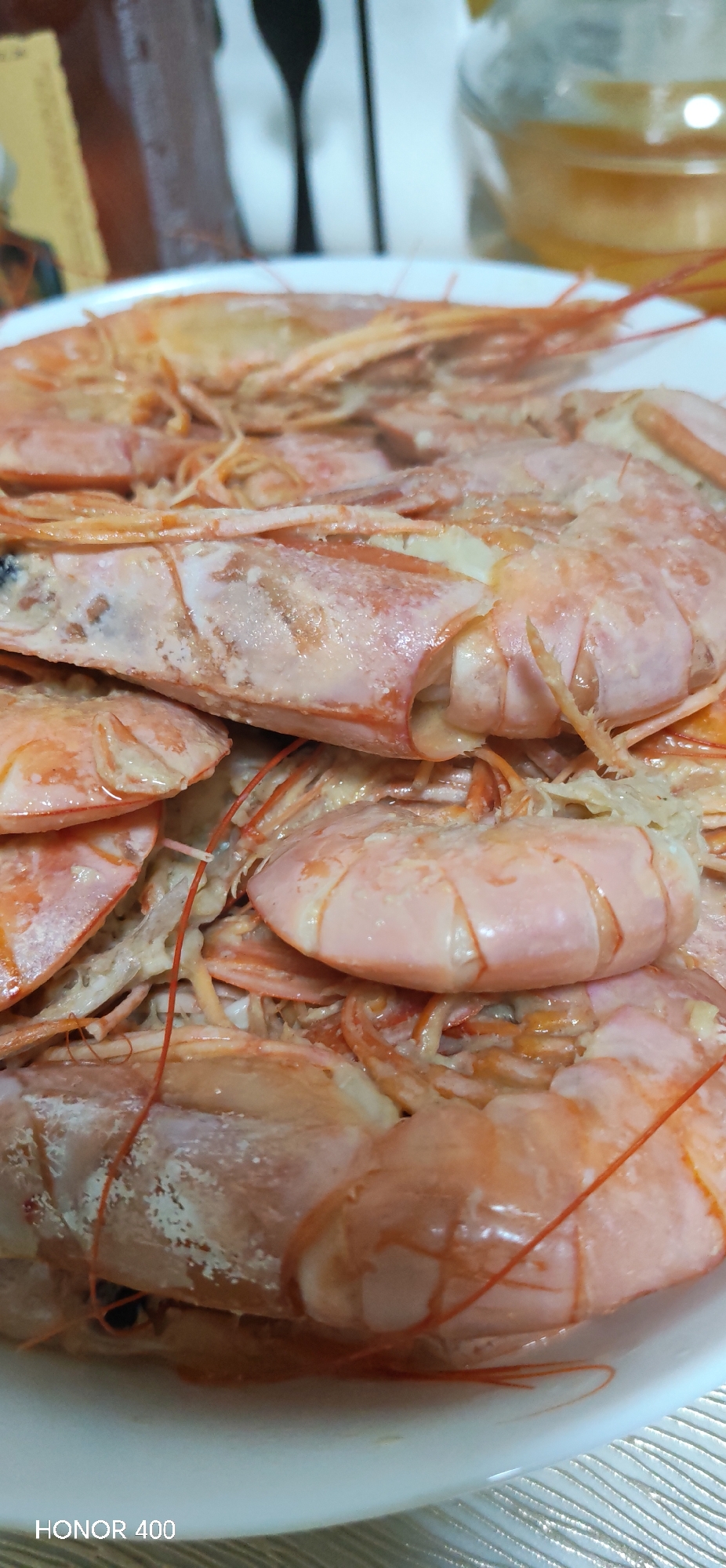 The image displays a close-up, top-down view of a serving of cooked shrimp or prawns. The subjects are piled within a white, shallow bowl or plate, which occupies the majority of the frame. The shrimp exhibit a reddish-orange to pale pink coloration, with some areas showing a lighter, almost white coating, possibly from salt or seasoning. Visible details include intact shells, antennae, and legs, with some internal white matter exposed.
In the blurred background, several objects are discernible:
To the upper left, a dark-brown, possibly glass bottle with a partially visible yellow label.
To the upper right, a transparent glass pitcher filled with a yellowish liquid.
Behind these items, a white wall or panel features a dark, stylized silhouette resembling a spoon or other kitchen utensil.
Below the white plate, a light-colored, textured surface, likely a placemat or tablecloth, displays a repeating pattern of diagonal ridges.
The scene is indoors, consistent with a dining or kitchen environment. No individuals are present or discernable, and no actions or interactions are depicted. The bottom left corner of the image contains a "HONOR 400" watermark.