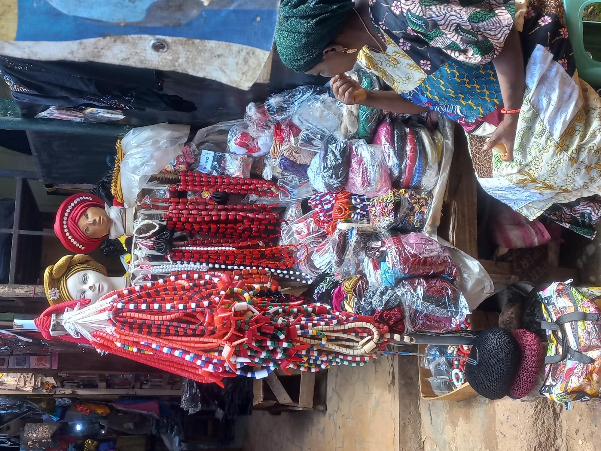 In a market stall in Jos, Nigeria, a woman with a green headscarf and a colorful, patchwork dress sits on a green stool, her right hand near her mouth. She faces away from the camera, towards a display of brightly colored necklaces and hair accessories. Two mannequins, one with a yellow headwrap and the other with a red one, are positioned behind the necklaces. The stall is filled with packaged items and hanging beaded jewelry, predominantly red and white, with some blue, green, and black accents. A patterned tote bag sits at the base of the stall, next to stacked netted hair accessories in black and maroon. The background shows stalls with more merchandise and textured walls.
