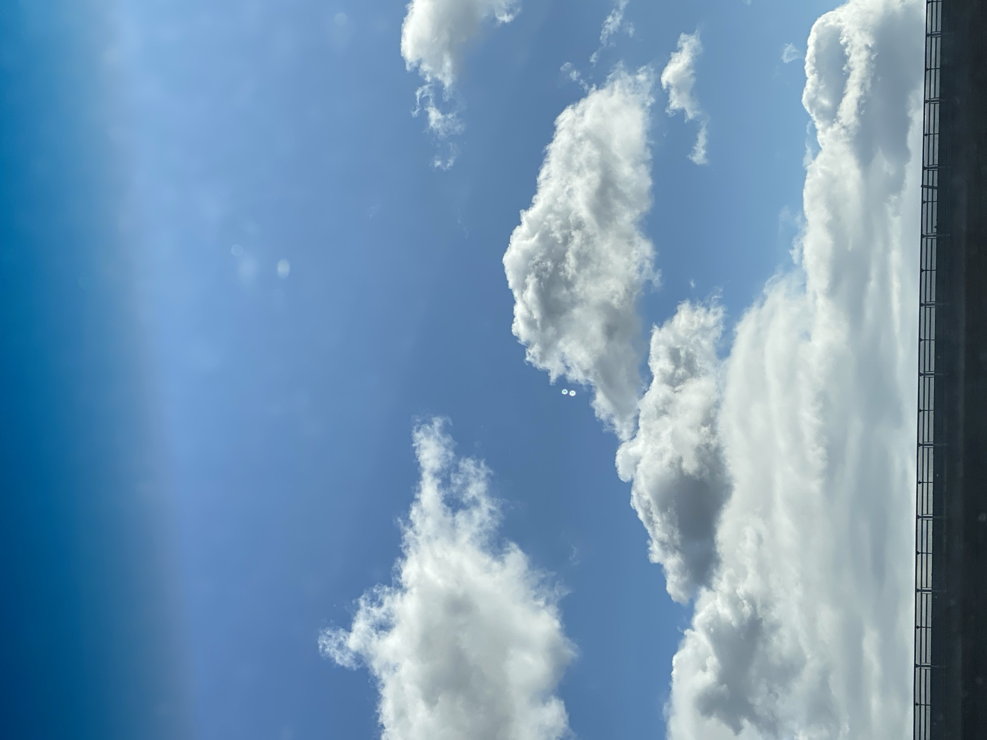 This image shows a bright blue sky with scattered white clouds above a dark-colored bridge railing. The railing spans horizontally across the lower portion of the image and appears to be made of metal with vertical supports and a horizontal top bar. The clouds are fluffy and voluminous, with some areas appearing brighter where they catch the sunlight. The sky transitions from a deeper blue at the top to a lighter blue near the horizon. There are no people or animals visible in the image. The scene suggests an outdoor environment, possibly a highway or elevated roadway, with a clear day. Based on the context, this scene could be located in Heringen/Helme, Germany.
