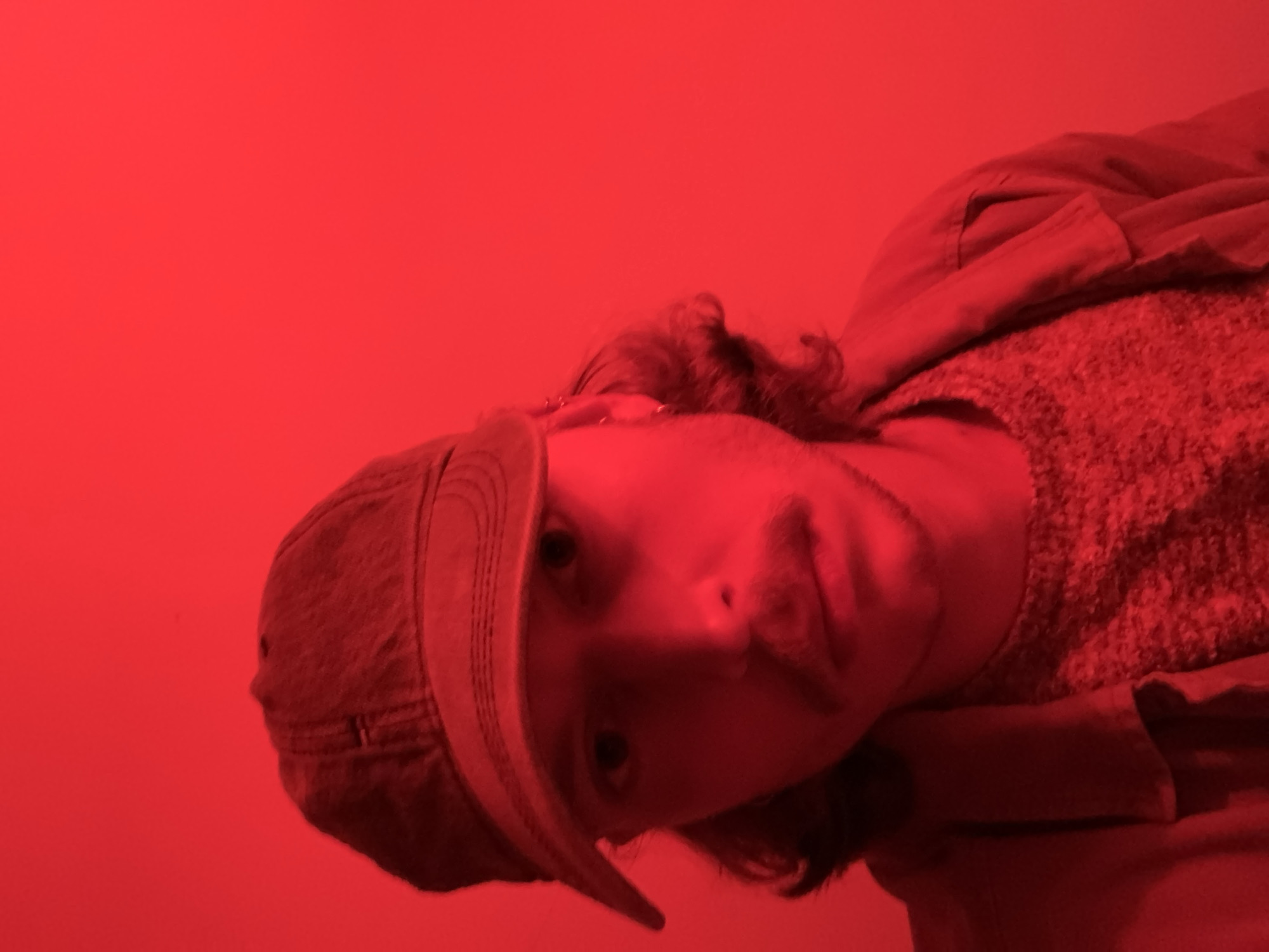 A person wearing a cap and jacket is illuminated by a strong red light. The background is a plain red color. It is not possible to determine the location or any details beyond what is directly visible in the image.