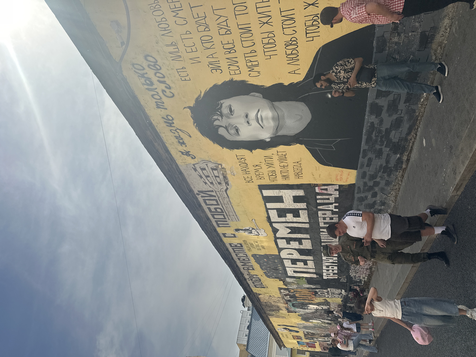 This image captures a vibrant street scene featuring the iconic Viktor Tsoi Wall, located on Krivoarbatsky Lane, near Arbat Street in Moscow, Russia, on a bright, sunny day. The central subject is a long, yellow-painted wall adorned with a large, black and white mural of Viktor Tsoi, the prominent Soviet rock musician, occupying the right-center portion. The wall is extensively covered with Cyrillic text, primarily lyrics from Tsoi's songs. To the right of Tsoi's portrait, lyrics such as "Я жизнь только слово" (My life is just a word) and "Есть лишь любовь и есть смерть..." (There is only love and there is death...) are visible. To the left of Tsoi, text reads "Все находят время, чтобы уйти, никто не уйдёт навсегда..." (Everyone finds time to leave, no one will leave forever...). Further left, a large, dominant black block text spells "ПЕРЕМЕН!" (Changes!) with "ТРЕБУЮТ НАШИ СЕРДЦА" (Our hearts demand) below it. The upper left part of the wall also features "ПОЕМ ВМЕСТЕ С ТОБОЙ" (We sing with you). The bottom section of the wall is painted with a dark grey and black brick pattern. The building's roofline, dark and weathered, is visible along the top of the wall, with a clear blue sky showing scattered cirrus clouds and a strong sun glare in the upper right. Several individuals are walking or standing on the asphalt sidewalk in front of the mural. In the foreground left, a young girl, approximately 6-8 years old, with light brown hair in a ponytail, wears a white tank top, loose light-wash jeans, and white sneakers. She holds a pink item in her left hand and has her right hand near her face, possibly shielding her eyes from the sun or wiping them. She is walking away from the camera. In the mid-ground center, a man in a full olive-green military camouflage uniform, including a cap and dark boots, smiles while looking at another man beside him. This second man, with short dark hair, wears a white short-sleeved t-shirt with two black stripes on the sleeves, dark shorts, white athletic socks, and dark sneakers. He is looking at and holding a white smartphone in his left hand, also smiling. They appear to be conversing while walking. Further right, a man with a dark beard and sunglasses, dressed in a patterned short-sleeved shirt (light brown, dark brown, and black), blue jeans, and dark blue slip-on shoes, stands