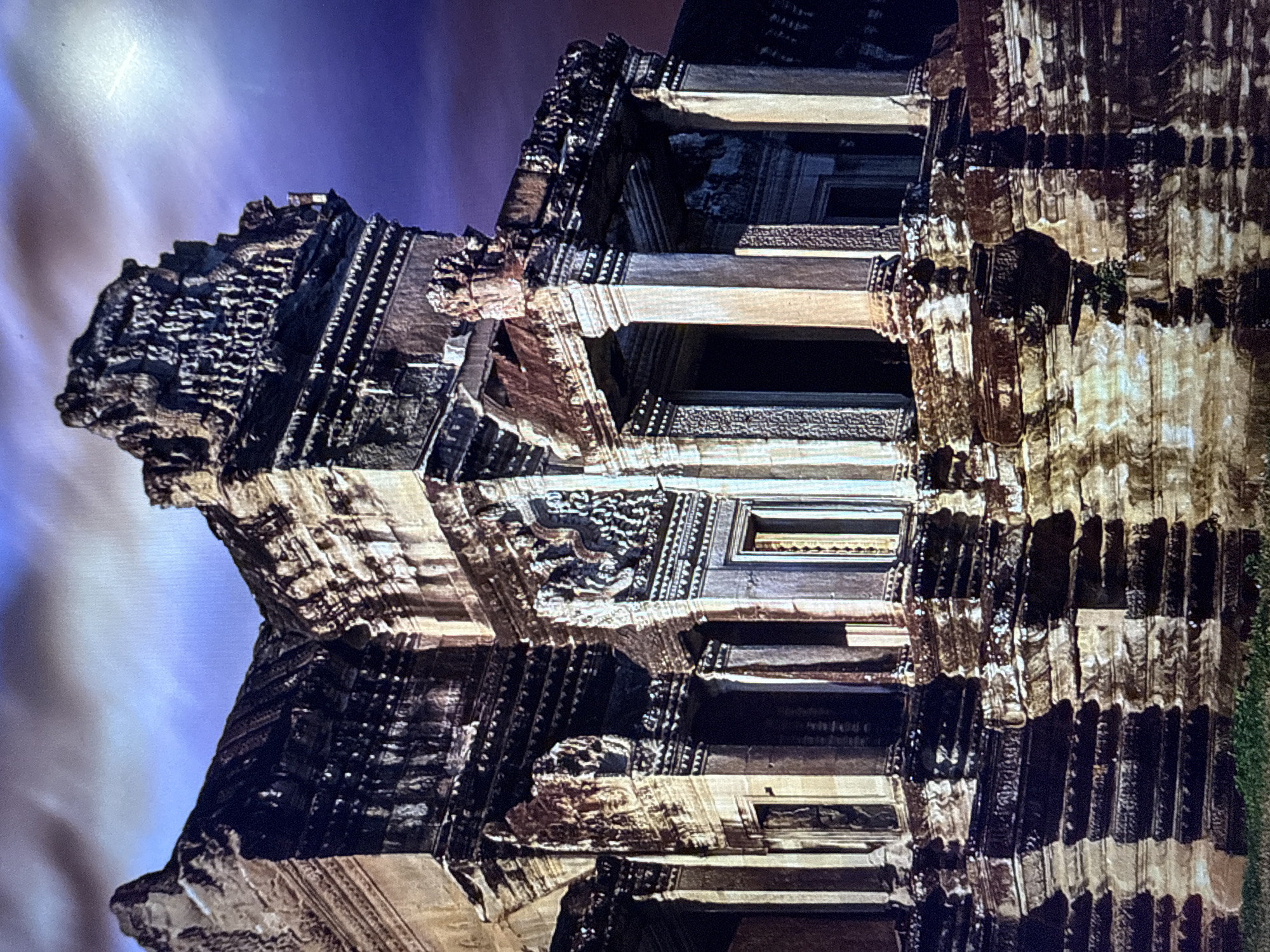 The image depicts the exterior of a large, ancient stone temple with intricate carvings and tiered structures. The architecture suggests Southeast Asian influence, possibly Khmer. The stone appears weathered, with some sections dark and others illuminated by dramatic lighting, casting deep shadows. The sky above is a deep purple with streaks of cloud, indicating dusk or dawn. The foreground shows hints of vegetation. The location provided, Urbanització Vescomtat De Cabanyes, Spain, is inconsistent with the architectural style of the temple, suggesting a potential misidentification of the location. There are no people or animals visible in the image.