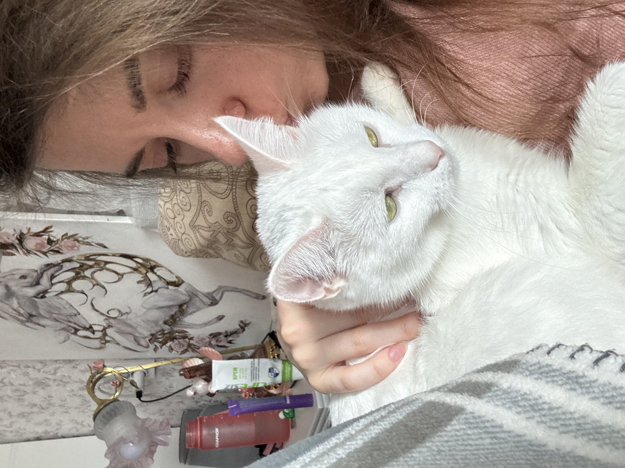 An indoor scene in the city of Chaikovsky, Russia, depicts a person affectionately kissing a white cat. The individual, seen from the upper torso, has light brown hair and is wearing a reddish-brown garment. Their eyes are closed as they lean forward to kiss the cat's head/ear. The cat is entirely white with striking greenish-yellow eyes and appears calm, looking slightly downwards. The person's left hand gently rests on the cat's back. To the left, a light-colored nightstand holds a pink "AQUAPHOR" water bottle, a purple tube, a small green item, and a white tube of cream labeled "КРЕМ". A decorative lamp with a pinkish-white ruffled shade and a golden base, adorned with two small white bird figurines, stands on the nightstand. A dark cable is visible near the lamp. The wall features subtle patterned wallpaper and a prominent decorative piece depicting a white, horse-like creature with elaborate golden scrollwork and pink floral accents. A patterned pillow is partially visible behind the person's head. A light gray, textured blanket or throw partially covers the person and the cat.