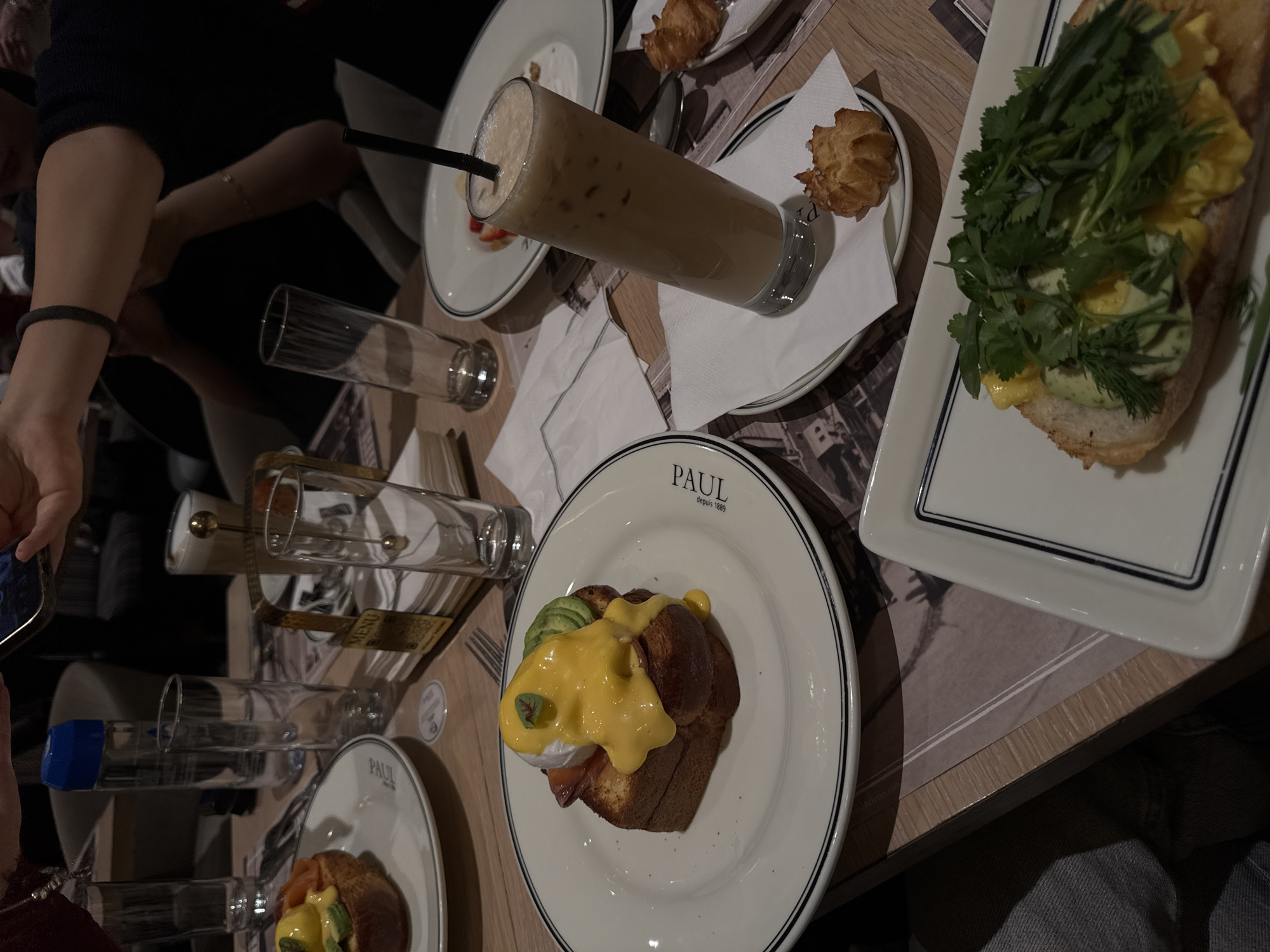 The image captures a dining scene at a PAUL cafe, identified by the distinctive "PAUL depuis 1889" branding on the plates, located in Yerevan, Armenia. The view is an overhead shot of a wooden table set for a meal.
Several food items are prominently displayed. A central white plate holds a portion of Eggs Benedict, featuring a poached egg, smoked salmon, avocado slices, and a rich yellow hollandaise sauce, all served on a piece of brioche bread. Another rectangular plate presents an open-faced sandwich, likely a baguette, topped with scrambled eggs, a green sauce (possibly avocado-based), and an abundance of fresh herbs like cilantro and dill. Small choux pastries, reminiscent of profiteroles or cream puffs, are scattered on side plates, and sliced strawberries are visible on one plate.
Drinks include a tall glass of iced coffee or latte with a straw, and multiple clear drinking glasses, some containing water (one next to a blue-capped water bottle/filter).
Several people are present, indicated by visible hands and arms; one person's hand holds a smartphone, while another arm, wearing a dark hair tie, rests on the table. The table itself is made of light-colored wood, partially covered by patterned paper placemats. White napkins and cutlery are also visible. A small stand displaying "MENU" and a QR code is on the table. The indoor lighting suggests a comfortable, possibly evening or dimly lit daytime atmosphere, typical of a cafe.