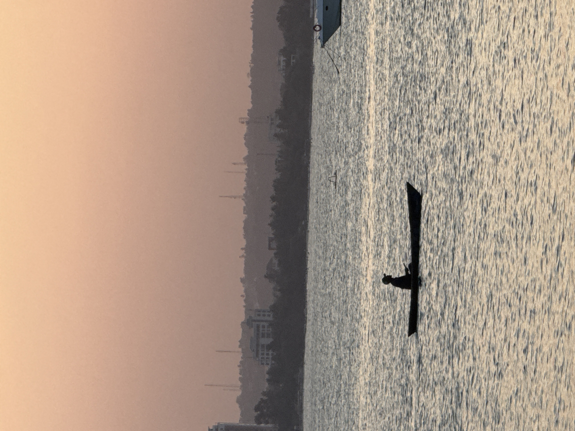 The image depicts a tranquil scene on the water, likely in Dar es Salaam, Tanzania, during the soft light of dawn or dusk. In the foreground, a lone figure is silhouetted in a small, dark boat, rowing across the gently rippling water. The figure appears to be wearing a hat. The water reflects the muted colors of the sky. In the mid-ground, a larger white boat is moored to the right. Further back, the coastline is visible, characterized by a dark silhouette of trees against a hazy sky. Several buildings, including a taller one and a larger, multi-story structure, are discernible within the treeline, suggesting an urban or semi-urban setting. Antennas or towers are also present on the horizon. The overall impression is one of peaceful solitude against a backdrop of encroaching development.