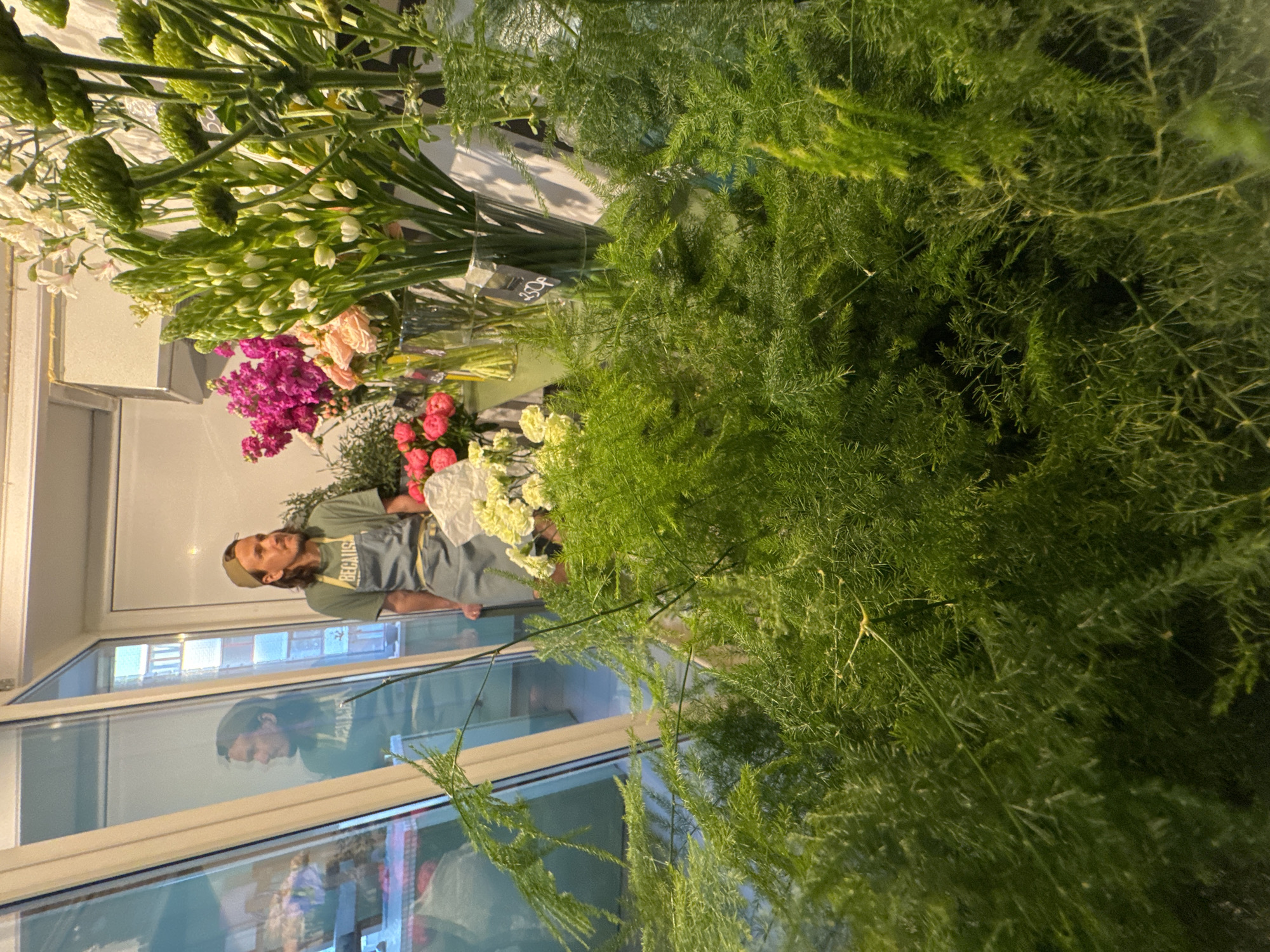 A man with long brown hair and a beard, wearing a green t-shirt and a gray apron, stands behind a display of flowers in what appears to be a florist shop. The flowers include white, green, pink, and red varieties. The display is positioned near a large window with reflections of buildings, suggesting an urban setting. Based on the information provided, this scene takes place in Izhevsk, Russia.