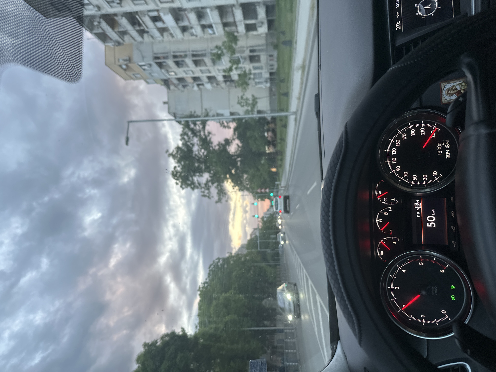 The image captures a view from inside a car, likely taken by the driver. The dashboard is prominent, showing a speedometer reading 50 km/h. The odometer reads 304164. The exterior scene features an urban environment with apartment buildings, streetlights, trees, and other vehicles on the road. The sky is cloudy and partially illuminated by the setting sun.
Based on the architecture and road layout, this location is potentially in the city of Plovdiv, Bulgaria. However, confirmation would require additional information such as street signs or landmarks.