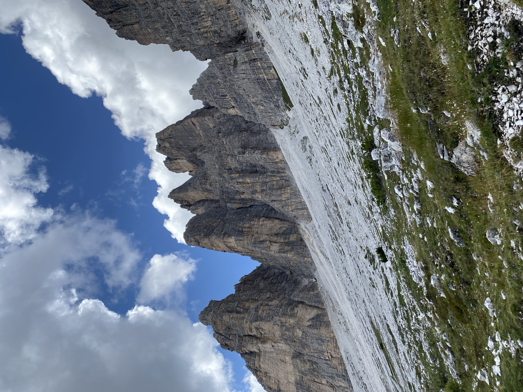 The image depicts a mountainous landscape under a partly cloudy sky. The foreground shows a grassy slope dotted with rocks and some sparse vegetation. The midground is dominated by a scree slope composed of light-colored rocks, ascending towards rugged, gray mountain peaks. The mountain peaks are tall, jagged, and appear to be made of stratified rock, with visible horizontal lines and vertical fissures. Some of the peaks are sharp and pointed, while others have more complex, fragmented summits. The sky above is a vibrant blue, with fluffy white clouds scattered across it, some of which are large and casting shadows on the mountains. The scene is devoid of any human presence or structures. The location is identified as being near Auronzo di Cadore, Italy, a region known for its dramatic Dolomite mountain scenery.