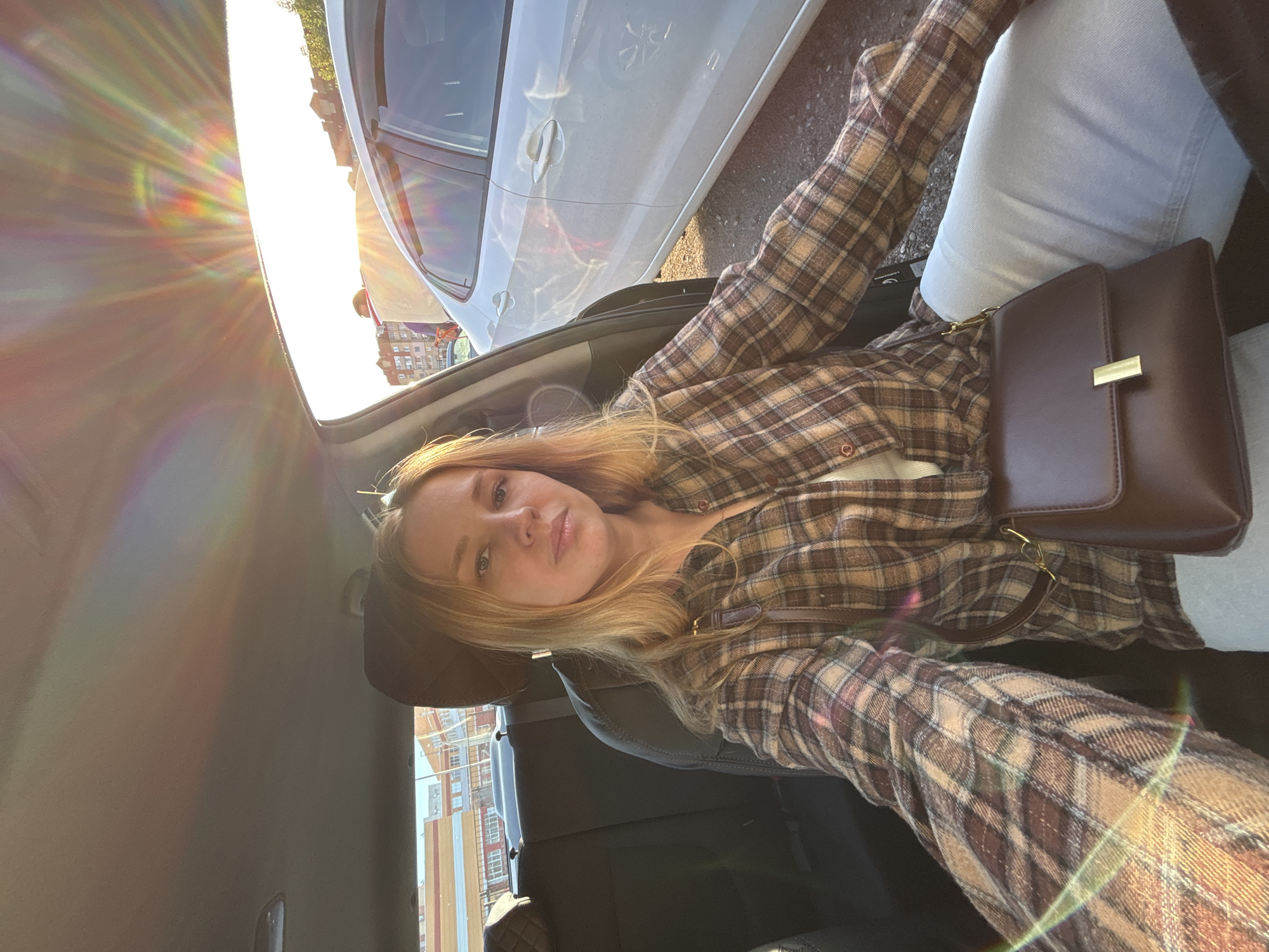 A young woman is captured in a selfie from inside a car, looking directly at the camera with a neutral expression. She has light-colored hair and is wearing a brown and beige plaid long-sleeved shirt, paired with light-colored jeans. A dark brown handbag with a gold clasp rests on her lap.
The setting appears to be within a car, with the dark interior of the car seat and light grey ceiling visible. Through the windows, a white car is parked nearby, and in the background, there are buildings and distant trees, suggesting an urban or suburban environment.
The time of day is characterized by strong, low-angle sunlight, producing prominent sun flares and a rainbow-like spectrum on the left side of the image, indicating either early morning or late afternoon. The weather is sunny and clear. No visible text is present in the image. The provided location context is Rusanovo, Russia.