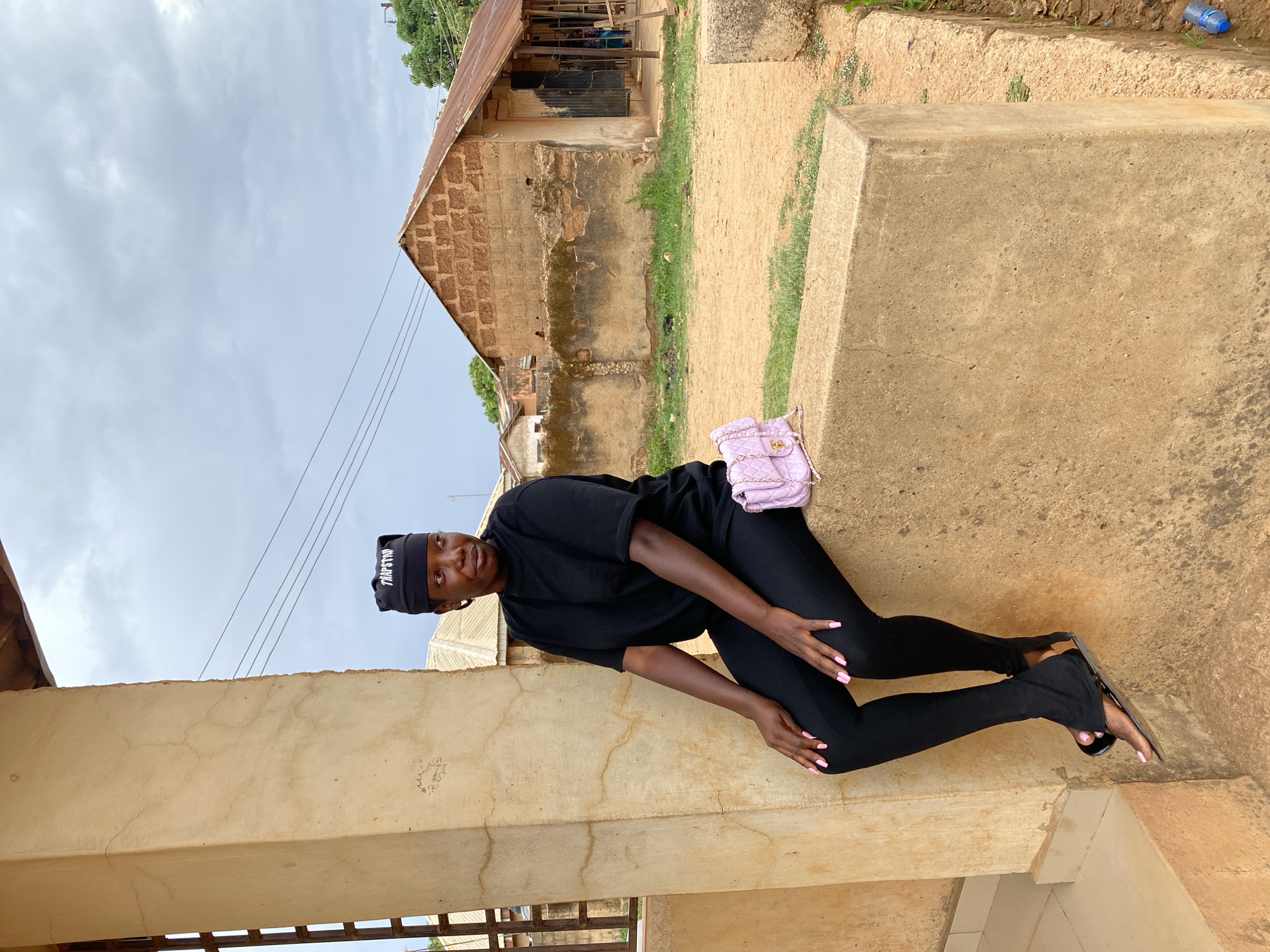 A young Black woman is depicted posing outdoors, leaning against a low, light-brown concrete structure. She is dressed in all black, wearing a black t-shirt, black leggings, and black open-toed sandals. A black beanie with the word "TRAPSTAR" written in white is on her head. A light pink quilted handbag with a chain strap rests on the concrete next to her. Her fingernails and toenails are painted with pink polish.
The background reveals a simple, possibly older building with textured, earthy-toned walls, some appearing to be exposed brick and others rendered. It has a rusted corrugated metal roof. The ground is mainly dirt with sparse patches of grass. Overhead, utility lines stretch across an overcast sky. The setting suggests a semi-rural or older urban area, consistent with Kuru Dungus, Nigeria. The woman is looking towards the camera with a calm, slightly smiling expression. The time of day appears to be daytime, with soft, diffused light indicating an overcast sky. A blue plastic bottle is faintly visible on the ground near a concrete step in one of the cropped images.