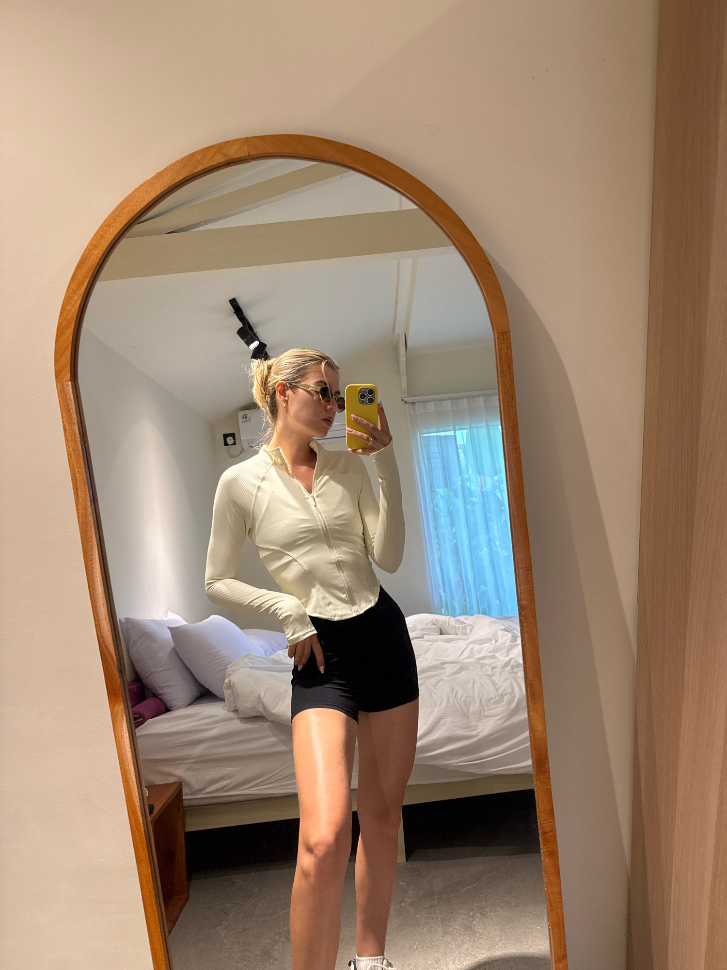 A woman is taking a selfie in a mirror in a room. She has blonde hair pulled back in a bun and is wearing sunglasses, a long-sleeved cream-colored top, and black shorts. The room appears to be a bedroom with a bed in the background covered in white linens and several pillows. There is a wooden nightstand next to the bed. The mirror is oval-shaped with a wooden frame and is leaning against a wall. A phone with a yellow case is visible in the woman's hand. The lighting is bright, suggesting it may be daytime.
