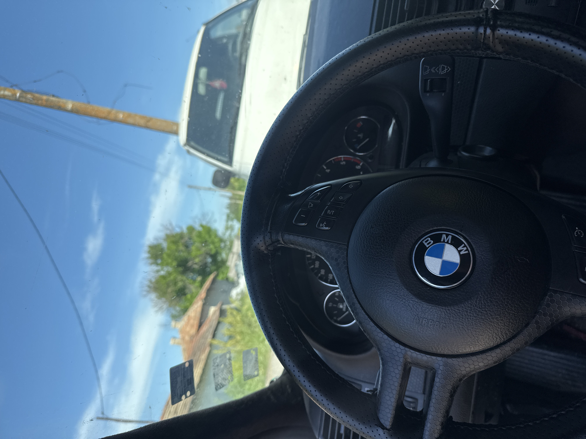 The image shows the interior of a BMW car, specifically the steering wheel with the BMW logo. The view through the windshield reveals a rural or semi-rural scene, likely in Alvanovo, Bulgaria. Visible outside are a white truck, a utility pole, a small building with a rusted roof, trees, and a clear blue sky. The steering wheel is black leather with perforated detailing, and buttons are located on the steering wheel. An airbag symbol is visible on the steering wheel hub. A windshield wiper lever is visible behind the steering wheel. The overall scene is captured from the driver's perspective.