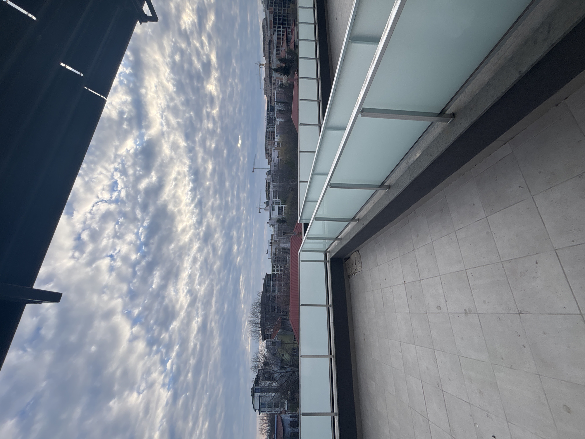 The image shows an elevated view from a balcony or similar structure. The floor is tiled with gray square tiles. A glass railing with metal supports runs along the edge of the balcony. Beyond the railing, the view opens up to a cityscape under a cloudy sky. Several buildings are visible, including residential blocks and structures with red rooftops. A crane can be seen in the distance. The cityscape appears to be Plovdiv, Bulgaria. To the left, a partial view of a dark structure is visible, possibly a building overhang. The sky is filled with textured clouds. There are no people visible in the image.