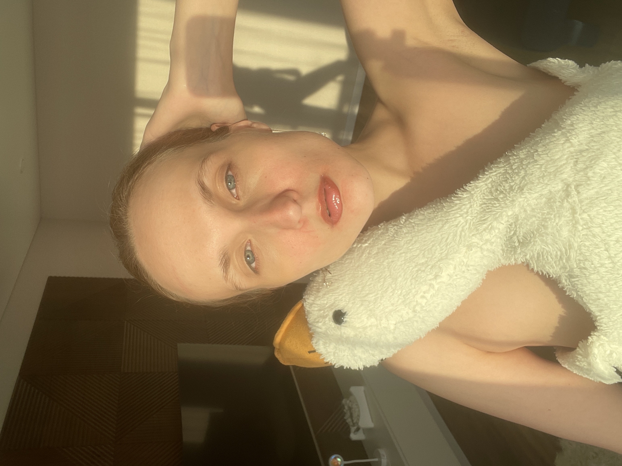 The image, taken in Podshivalovo, Russia, features a young woman indoors, bathed in natural sunlight. The photo is rotated 90 degrees counter-clockwise.
The central subject is a woman with fair skin, light-colored hair, and blue eyes. She looks directly at the camera with a calm, soft gaze. Her lips are glossy and slightly parted. She appears to be bare-chested, partially covered by a white, fluffy material, possibly a blanket or towel, draped over her torso and shoulder. Resting near her head and neck is a large, white, fluffy stuffed goose or duck with an orange beak and black eyes.
The setting is an indoor room. Strong, warm sunlight streams in from a window, creating distinct parallel shadows on the light-colored wall and the woman's skin, suggesting venetian blinds or a similar window covering. This lighting indicates either morning or late afternoon. The background also includes a dark, textured surface with a repeating geometric pattern, likely a piece of furniture like a headboard or a wall panel. A faint, blurred reflection or object can be seen on this dark surface.
The woman appears to be relaxing, possibly in bed or on a couch, and is posing for the photograph while holding or being accompanied by the stuffed animal. There is no visible text in the image.